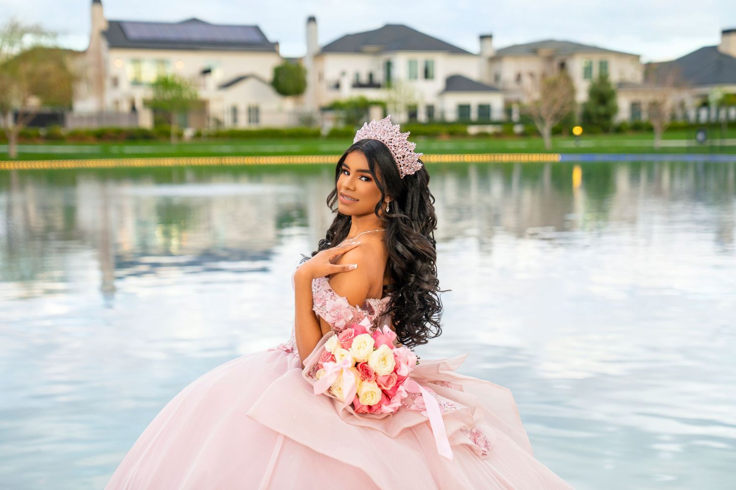 Young woman in pink quinceañera ball gown and floral crown holds bouquet by a lake with mansions in background.