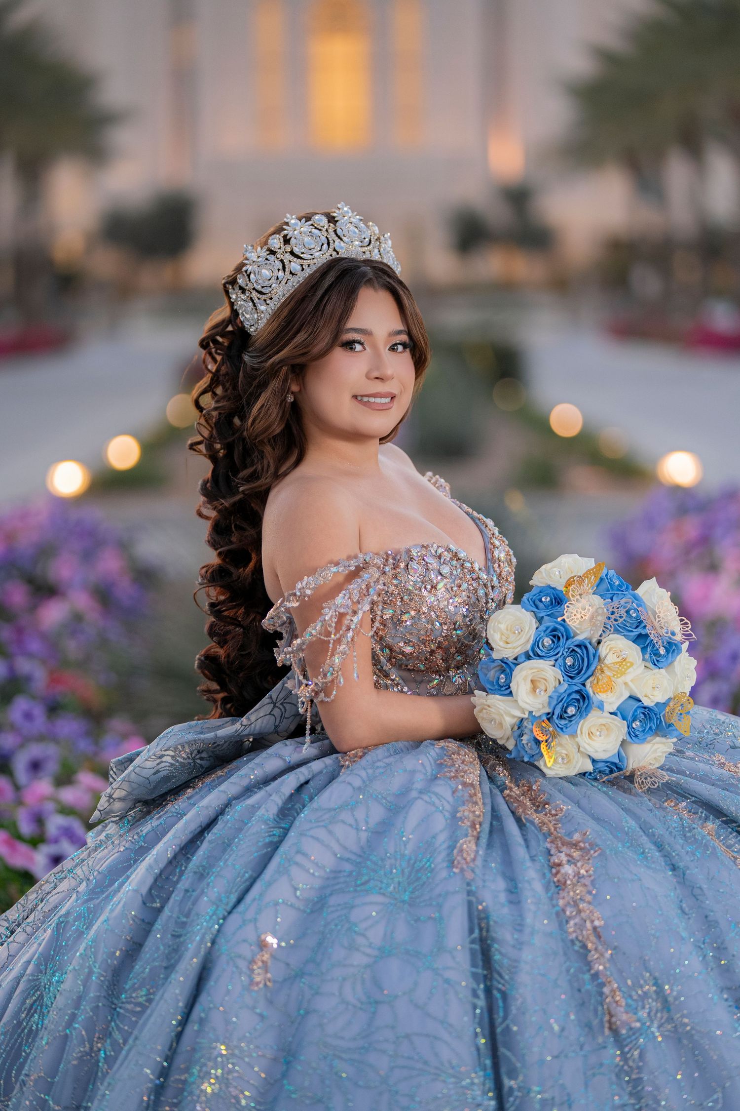 Young woman in a sparkling blue quinceañera ball gown and tiara holds a blue and white bouquet outdoors.