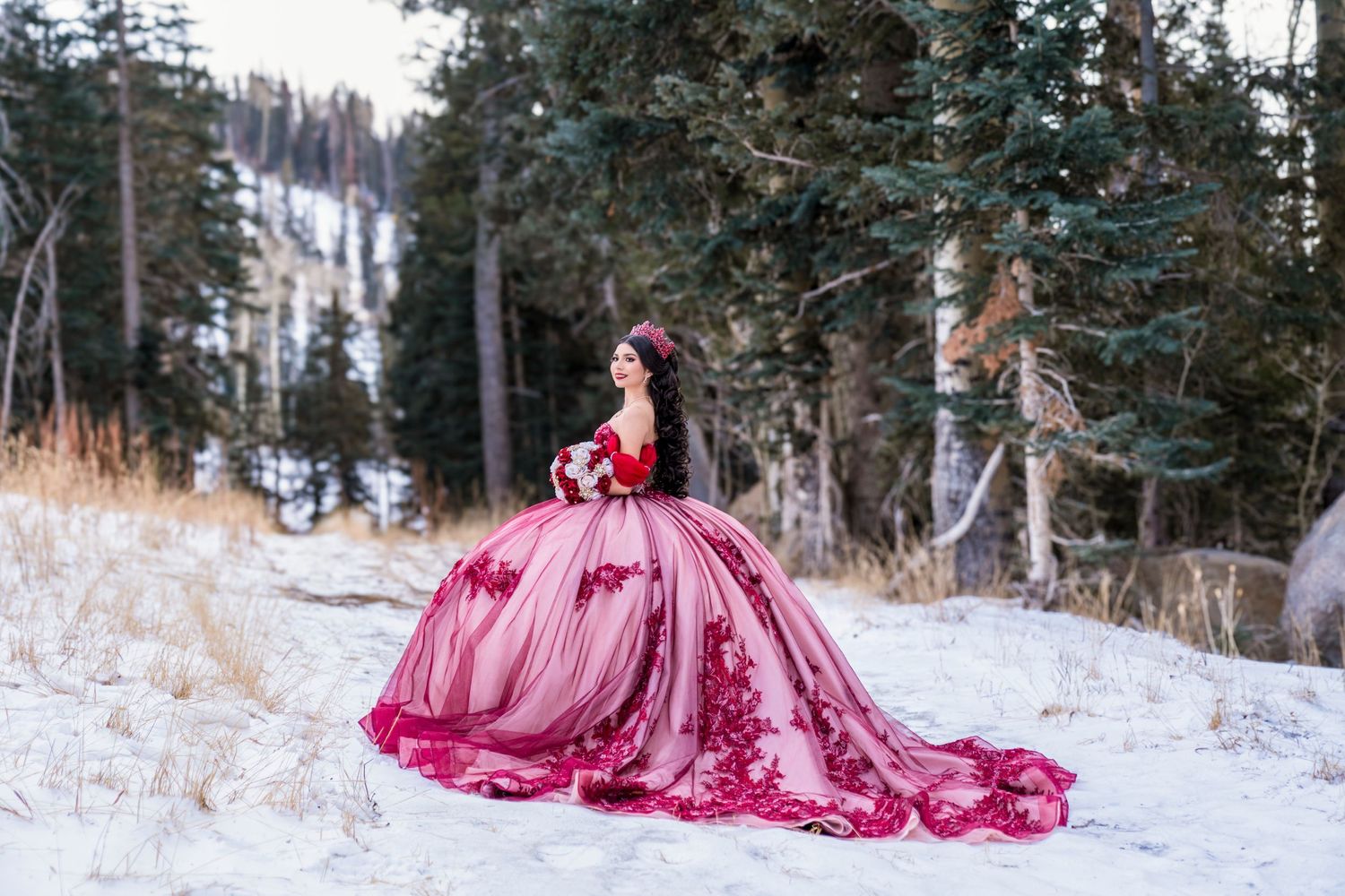 Woman in a vibrant red and pink quinceañera ball gown with long train stands in a snow-covered pine forest.