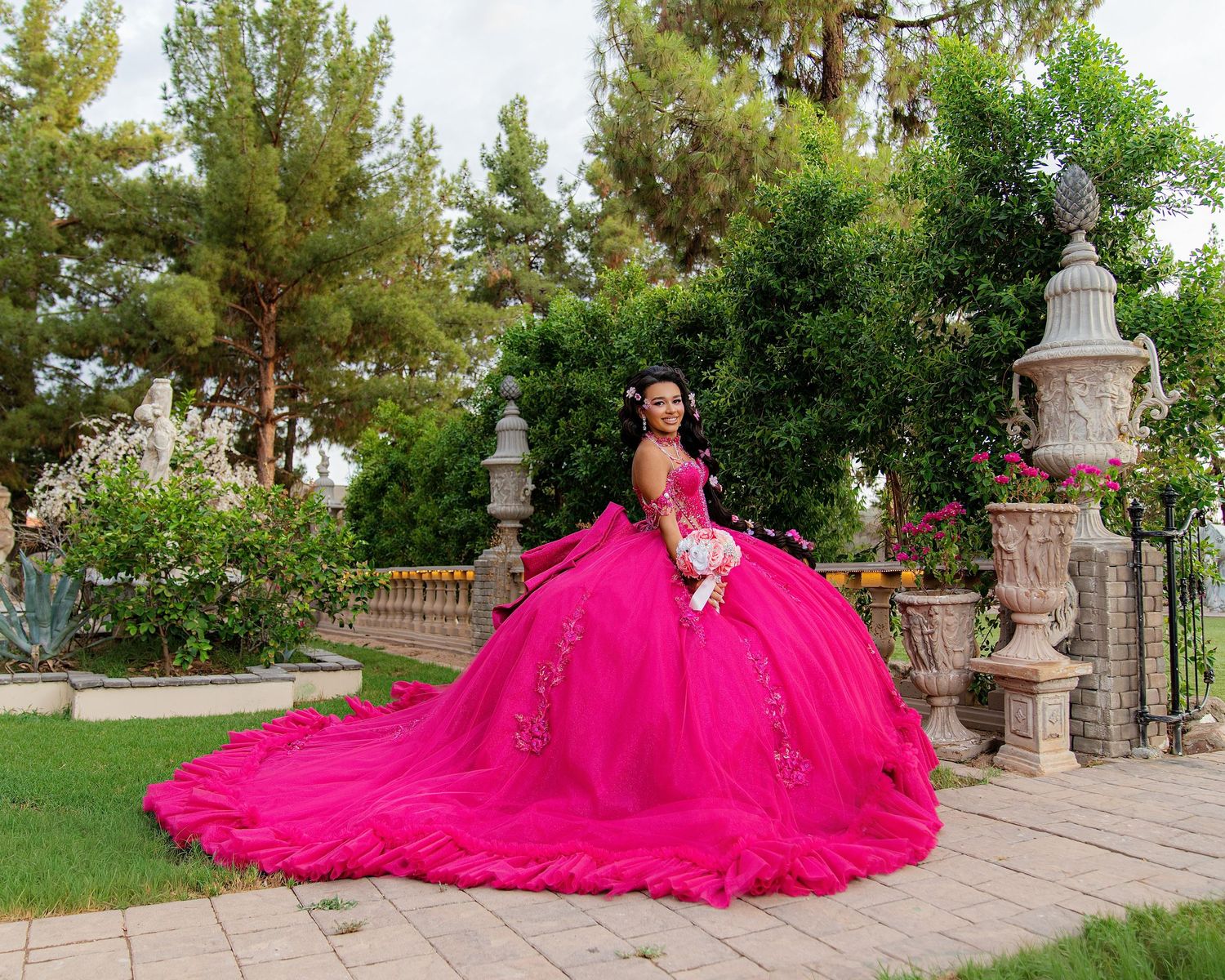 Young woman in a stunning hot pink ball gown quinceañera dress poses gracefully in a lush garden setting.