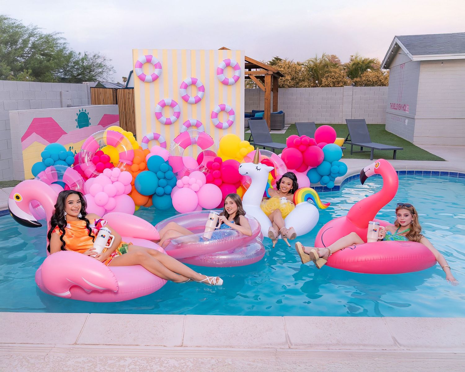 Four women enjoy a colorful pool party with flamingo floats, balloons, and festive decorations.