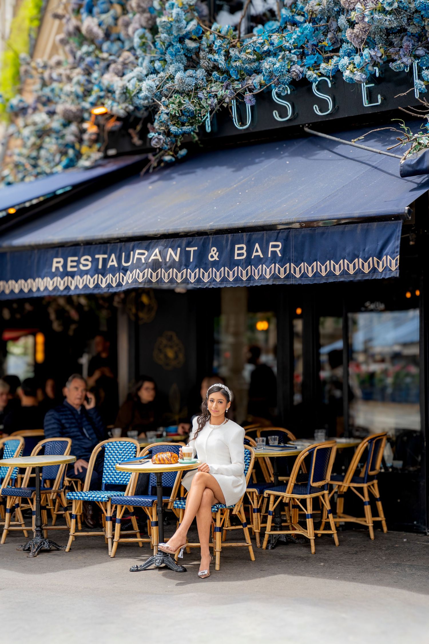 Stylish woman in white dress sits at outdoor cafe table beneath blue floral decor at a Paris restaurant.