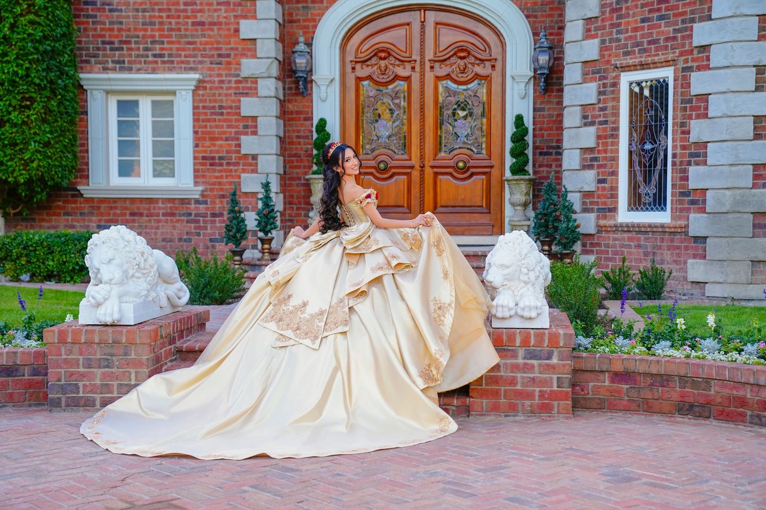 Elegant young woman in a gold quinceañera ball gown posing before a grand brick mansion entrance.