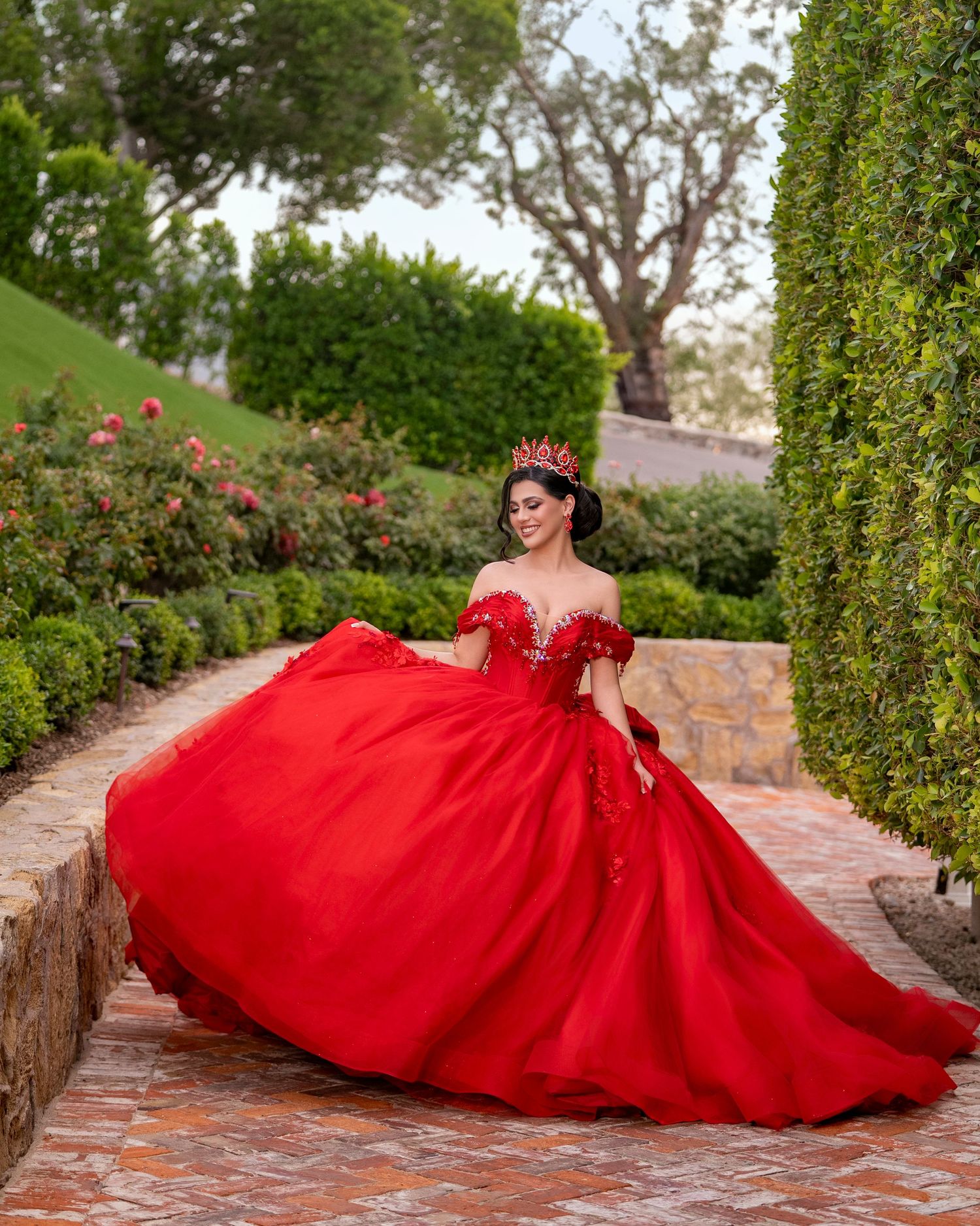 Young woman twirls in red quinceañera ball gown on brick garden path surrounded by lush greenery.