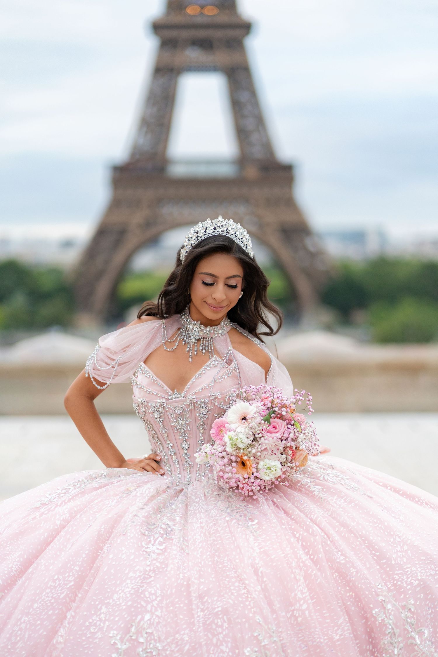 Young woman in pink ball gown and tiara holds bouquet with Eiffel Tower in background.