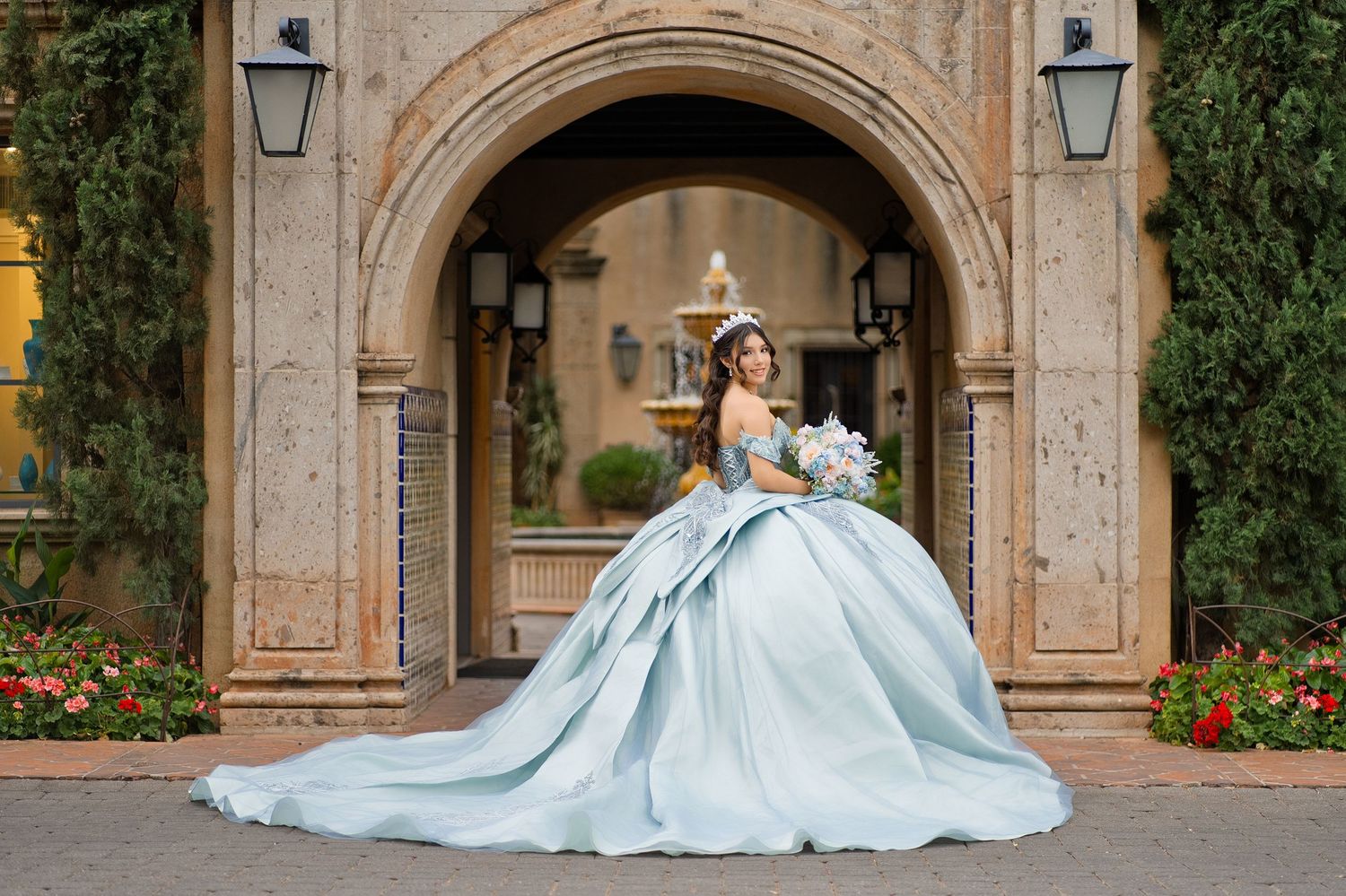 Young woman in a light blue ball gown quinceañera dress holding a bouquet, posing under an elegant stone archway.
