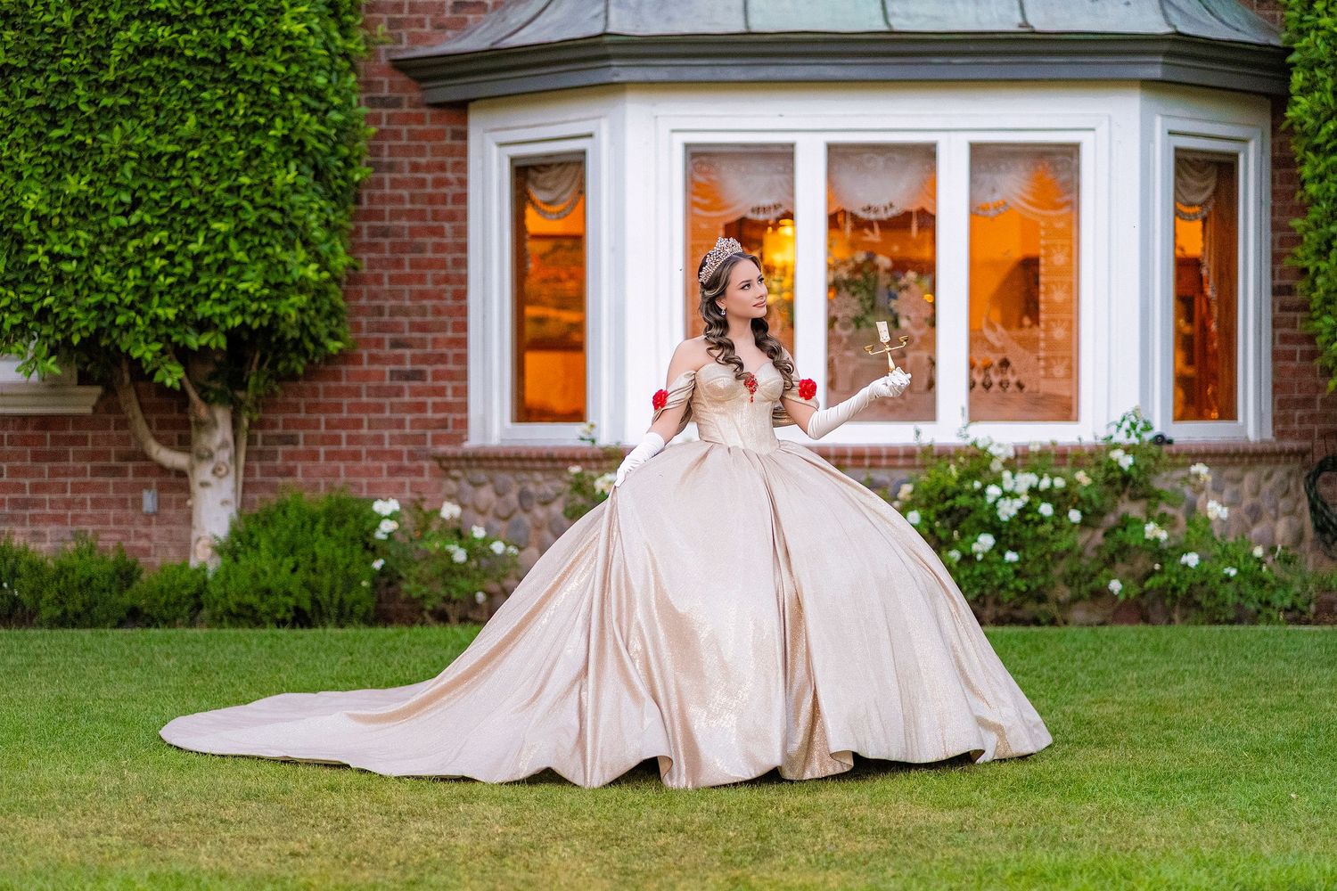 Woman in elegant champagne ball gown twirling on green lawn beside brick mansion with glowing bay window.
