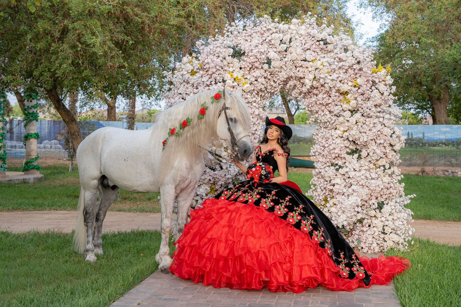 Girl in red and black quinceañera gown poses beside a white horse near a floral arch outdoors.