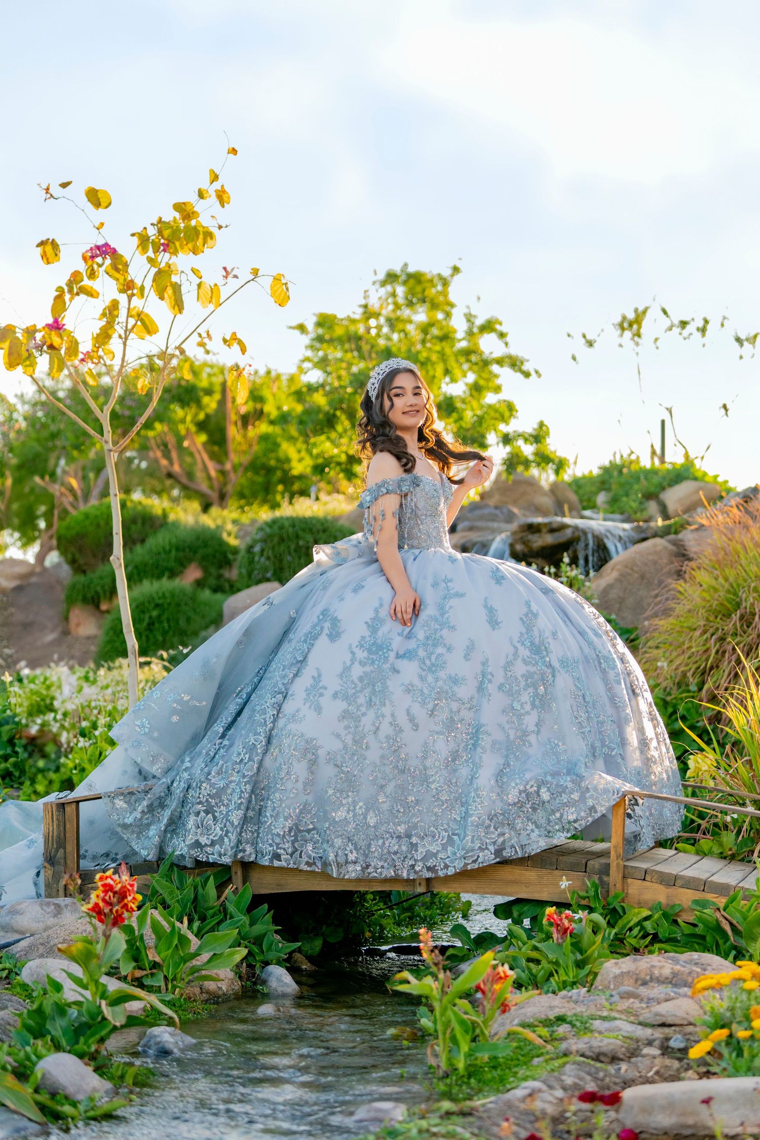 Young woman in a blue ball gown quinceañera dress poses on a garden bridge surrounded by lush greenery.