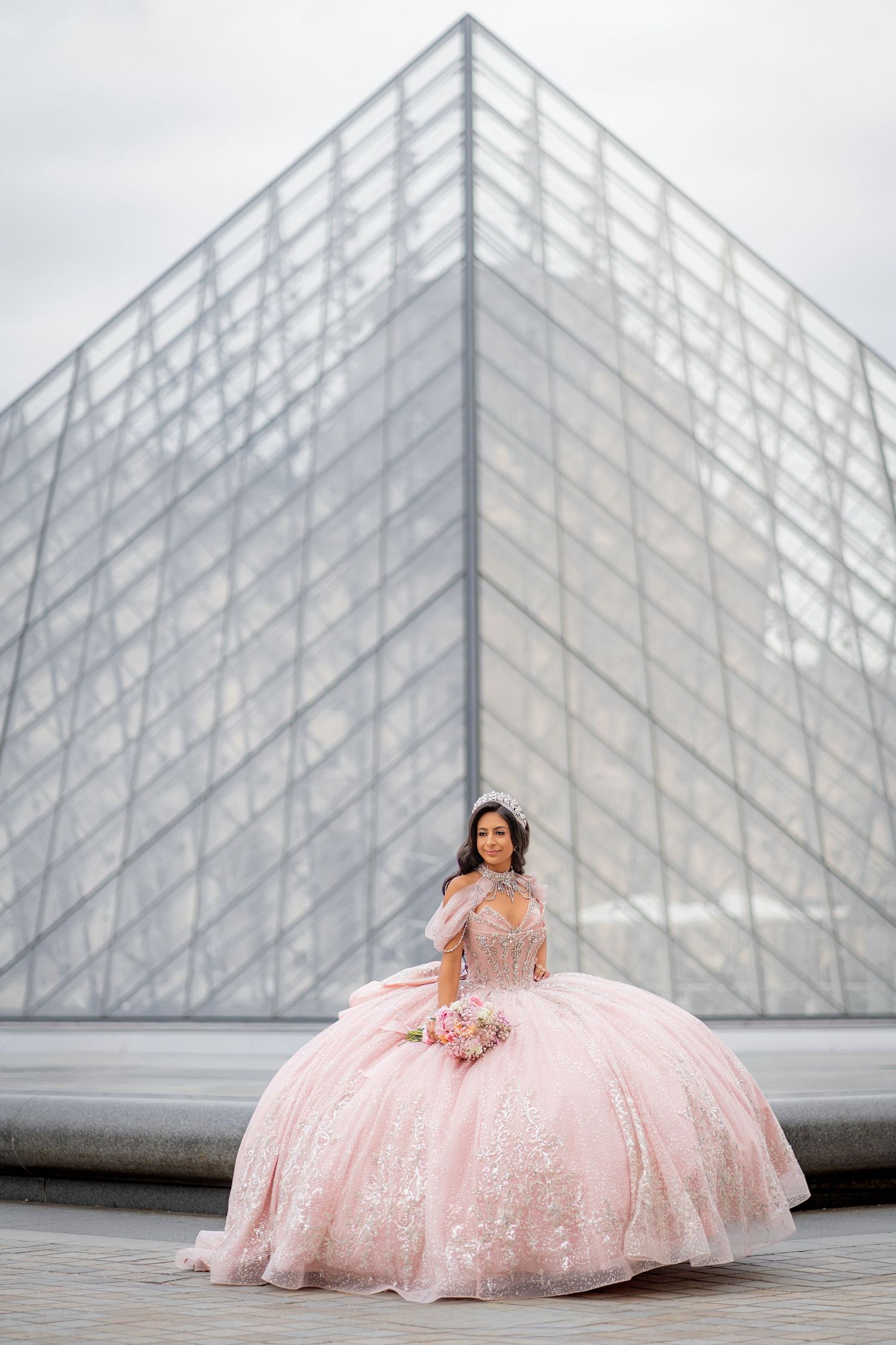 Young woman in pink quinceañera ball gown sits before the glass Louvre Pyramid in Paris.