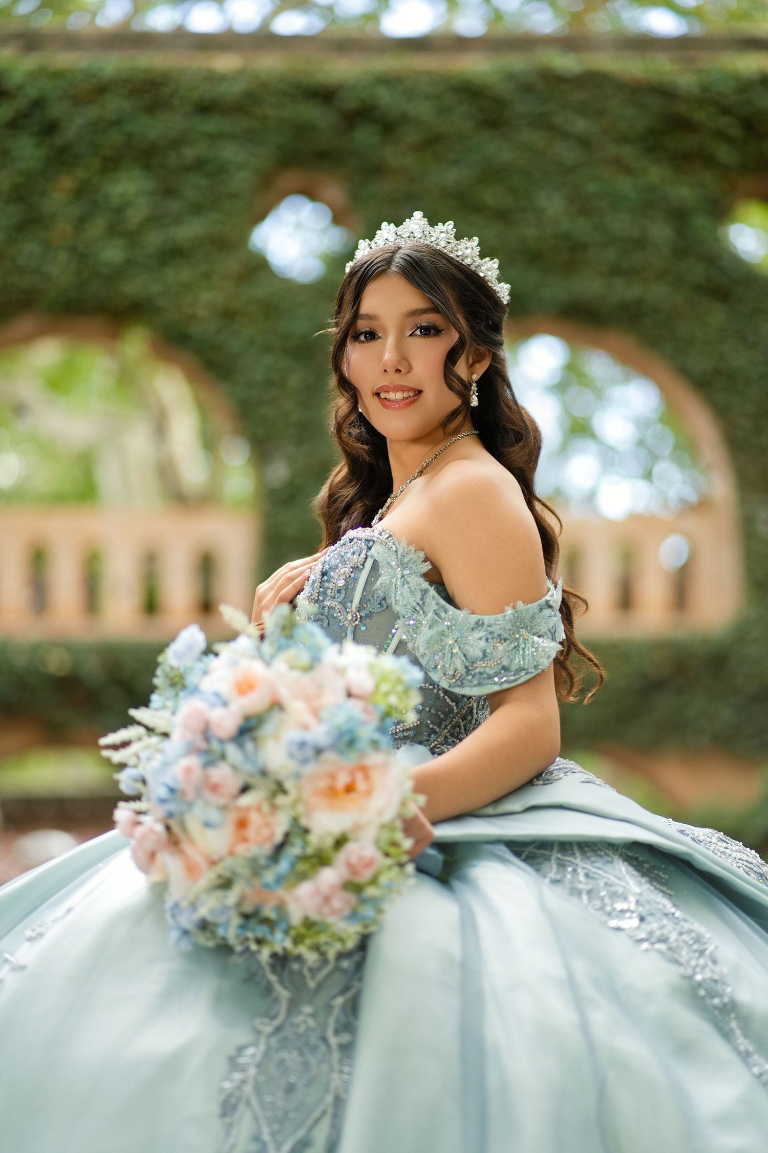 Young woman in blue quinceañera ball gown with tiara holds pastel bouquet in ivy-covered garden.