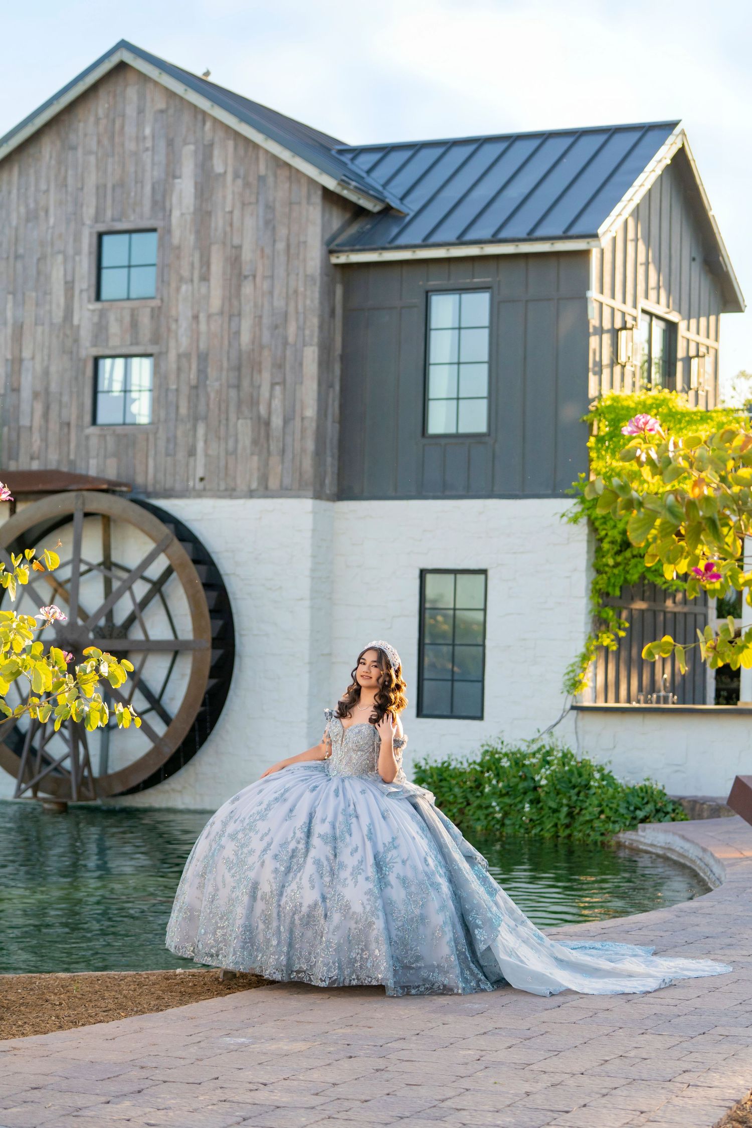 Young woman in elegant blue ball gown poses by a water wheel at a rustic venue.