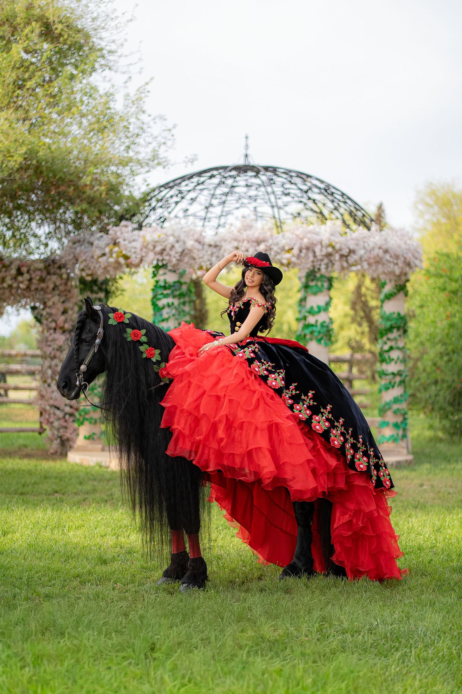 Young woman in red and black quinceañera gown on black horse near floral gazebo in garden.