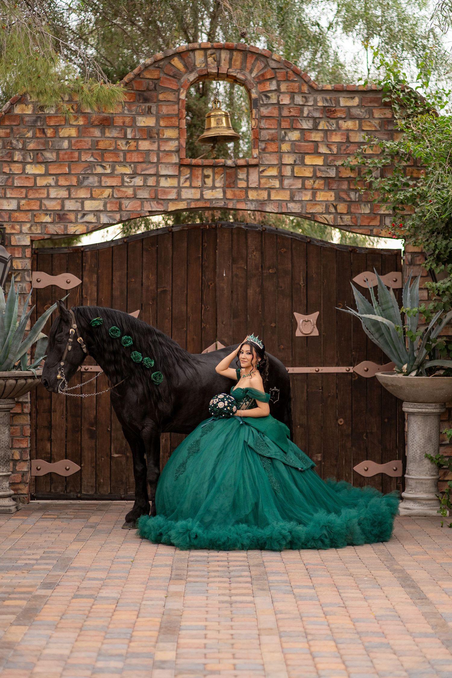 Young woman in emerald green quinceañera ball gown poses with black horse before rustic wooden gate with bell.