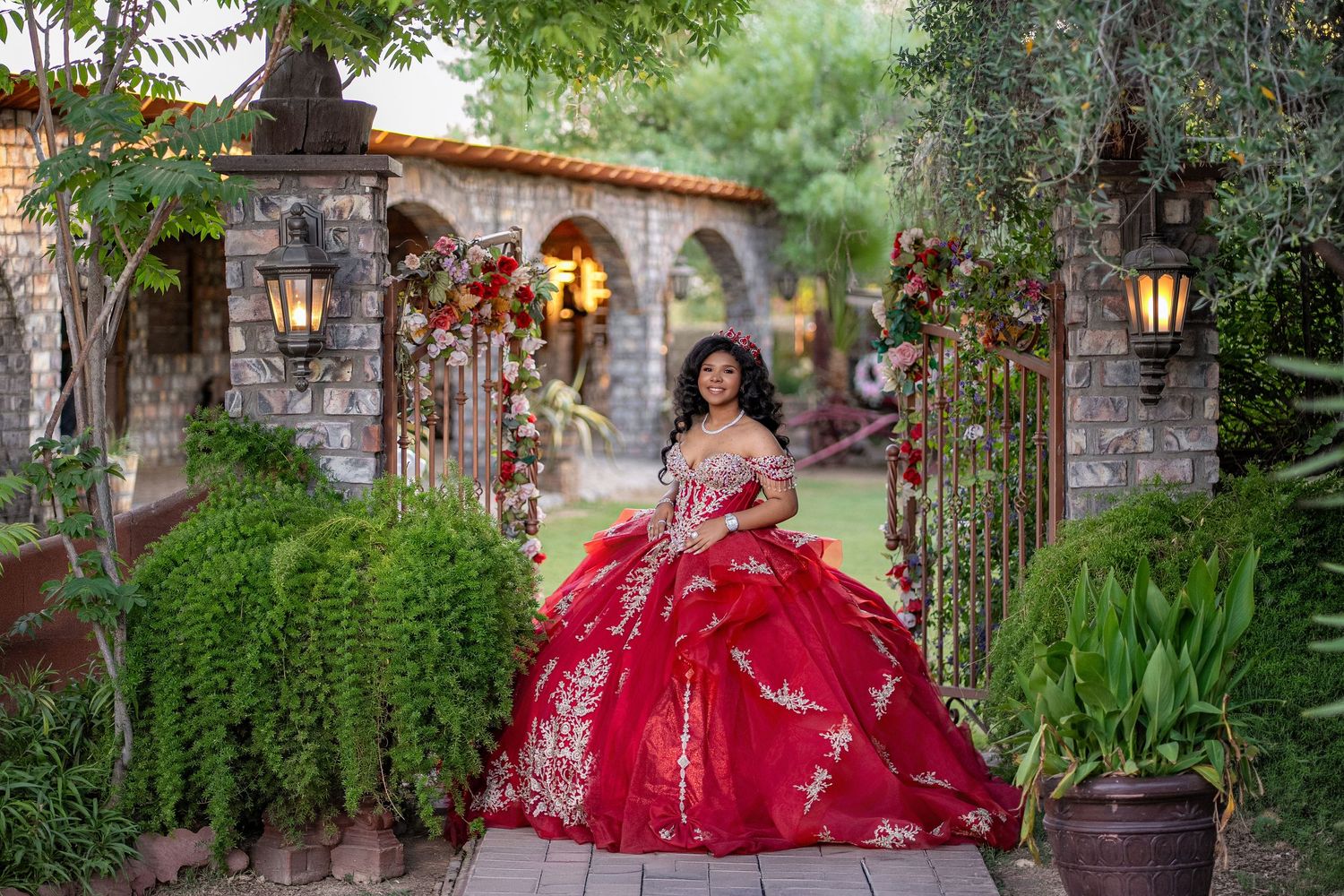 Young woman in red quinceañera ball gown with gold embroidery poses in lush garden with stone archway.