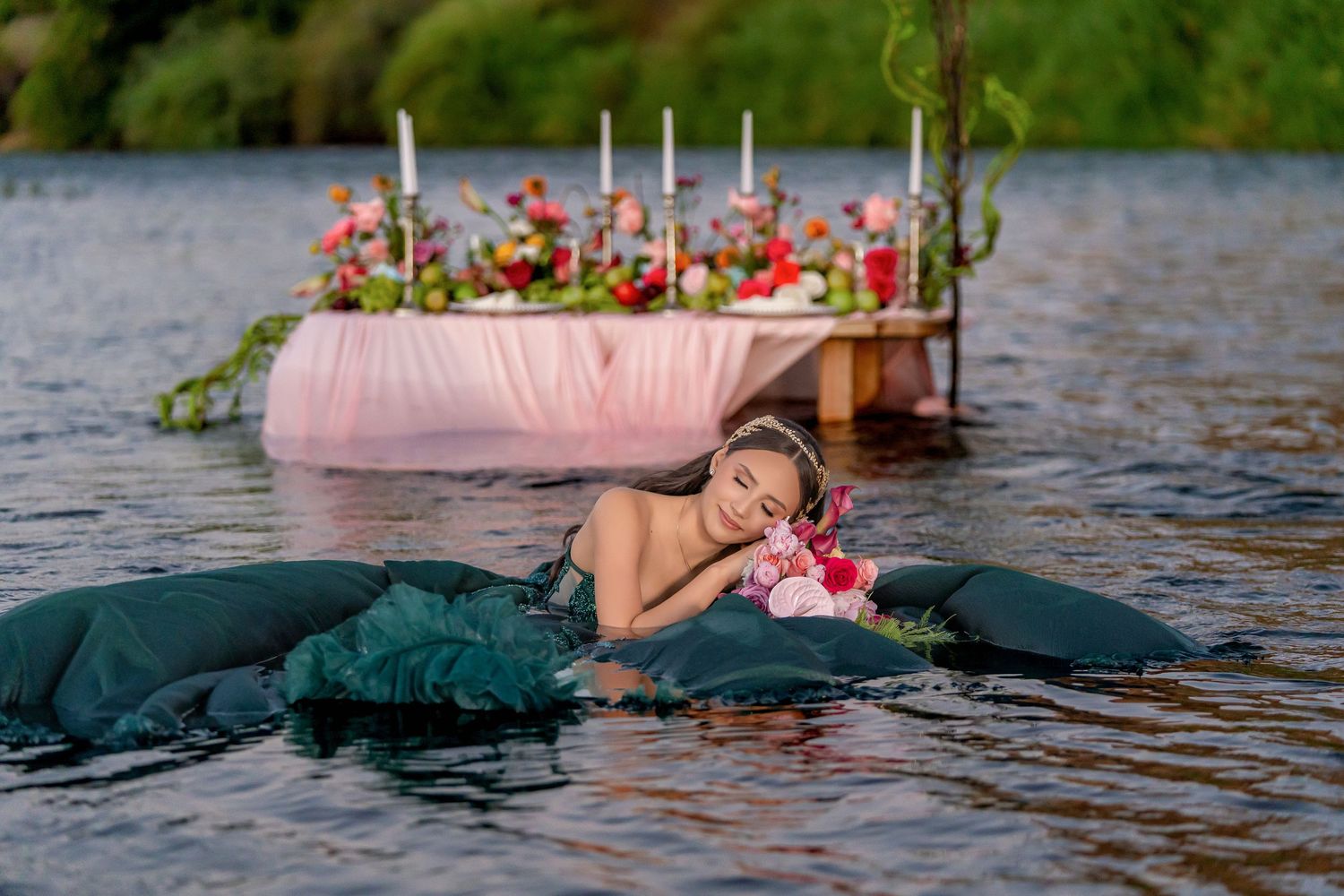 Woman in teal dress lying on float in lake with floral decorated table and candles in background.