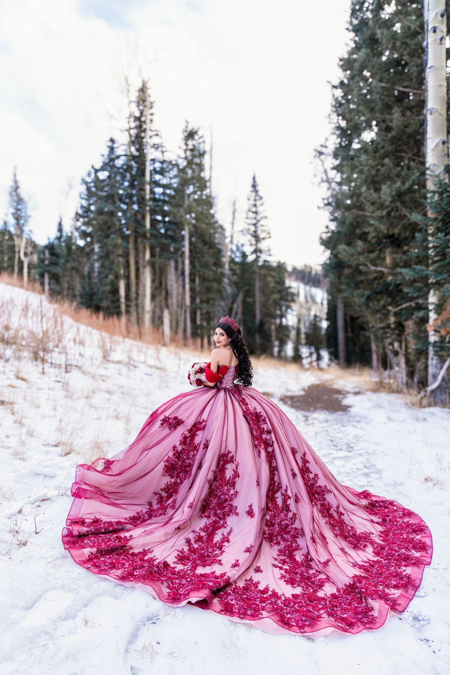 Young woman in a stunning pink and red floral quinceañera gown poses on a snowy forest trail.