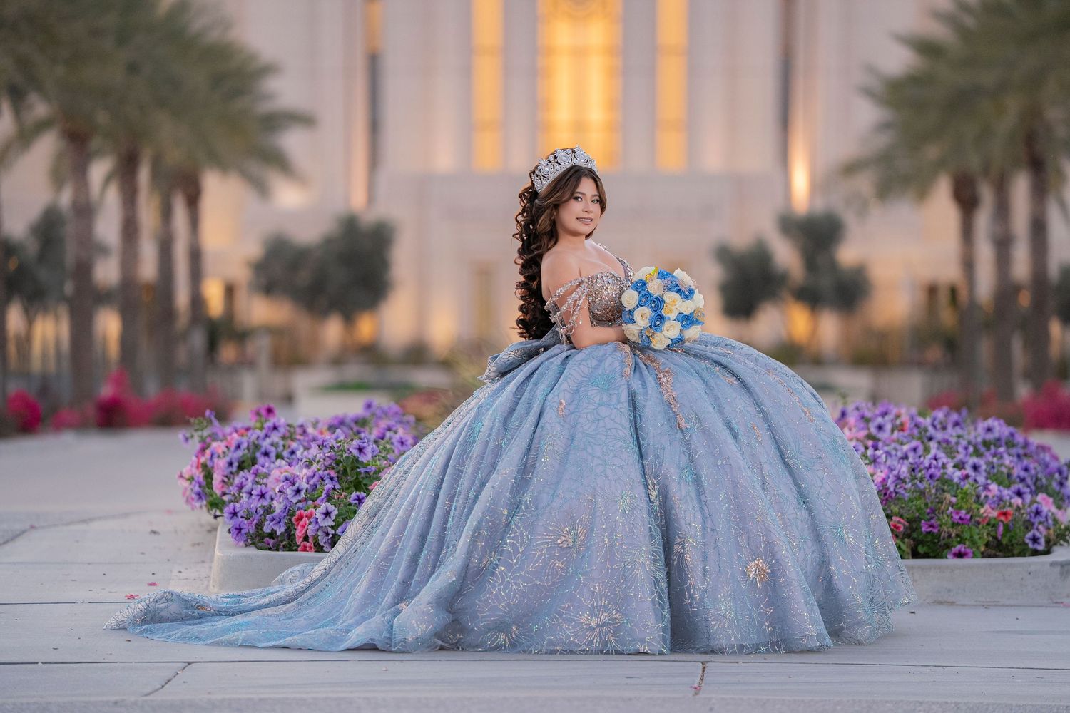 Young woman in blue quinceañera ballgown with floral crown holds bouquet beside colorful garden at elegant venue.