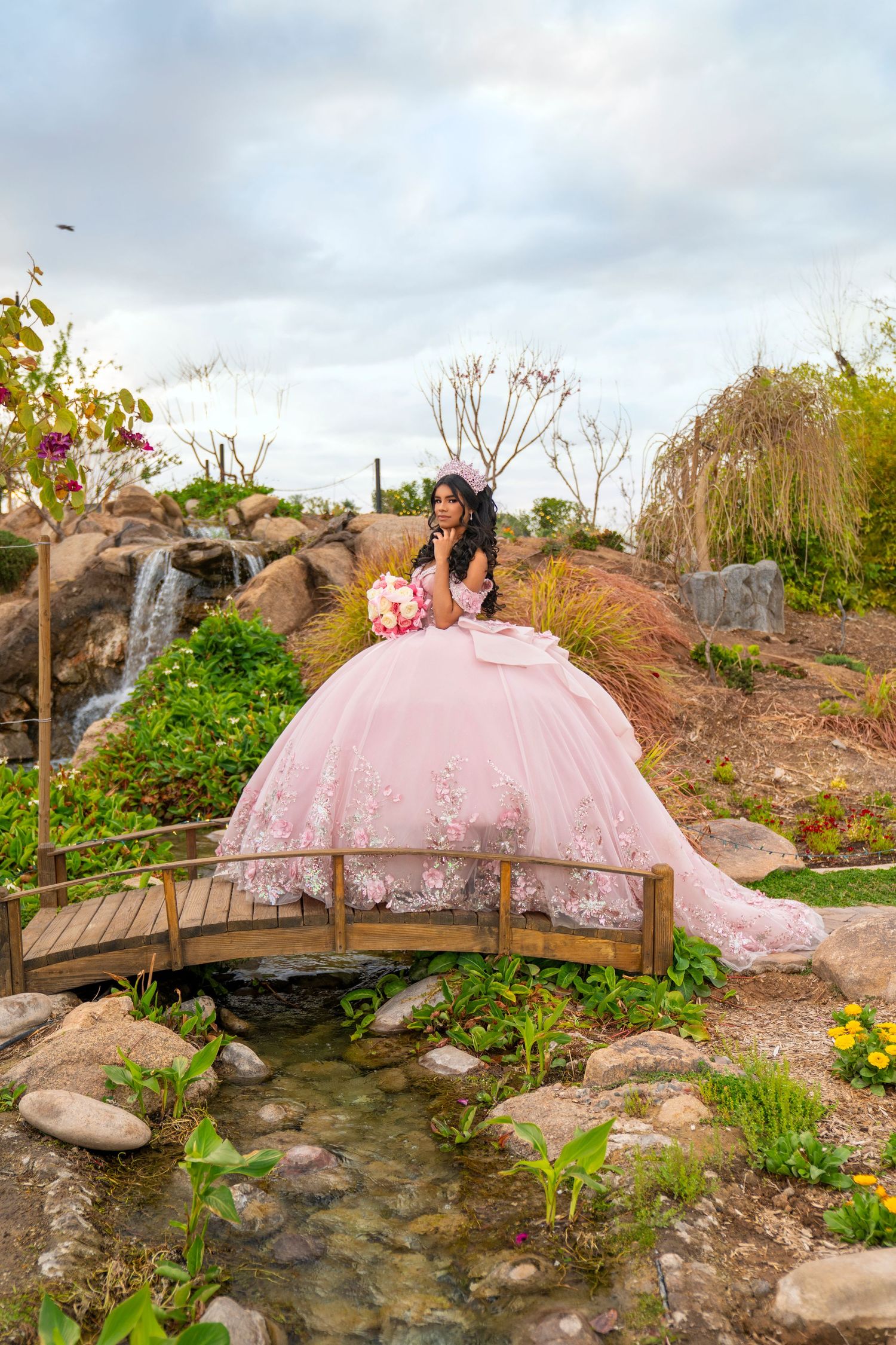 Young woman in a pink ball gown quinceañera dress poses on a wooden bridge surrounded by a lush garden waterfall.