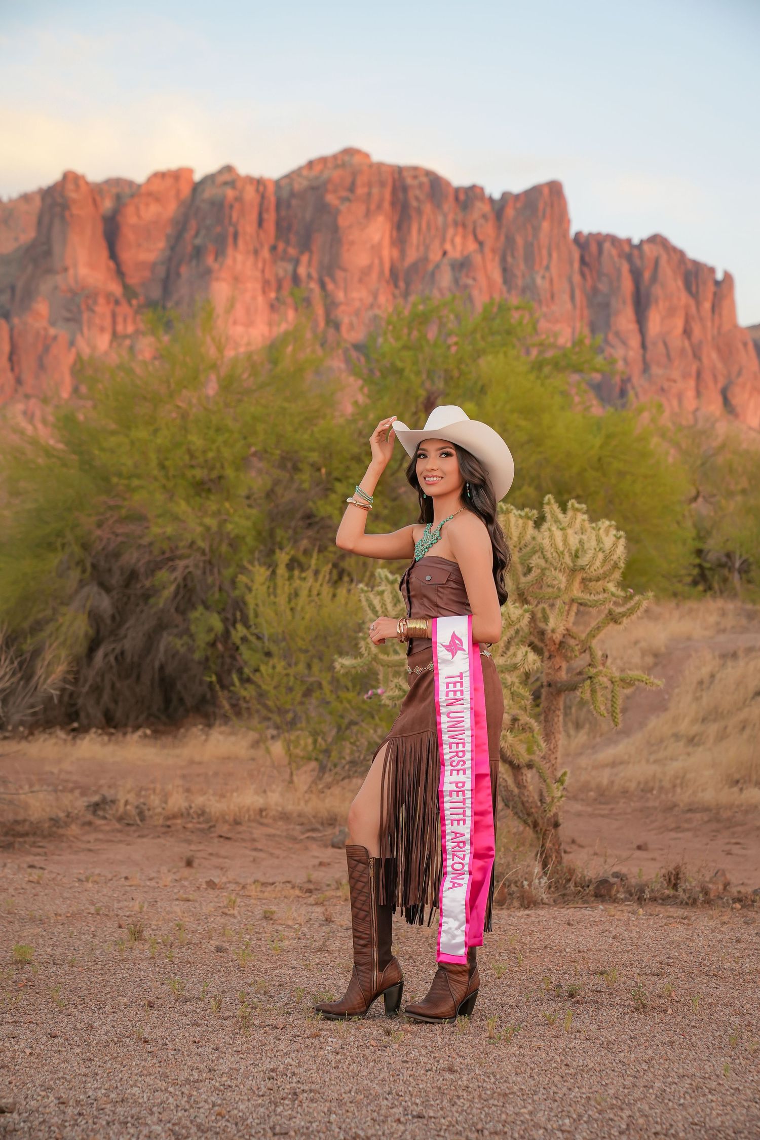 Woman in cowboy hat, brown fringe dress, boots, and pageant sash poses before red rock mountains in desert landscape.