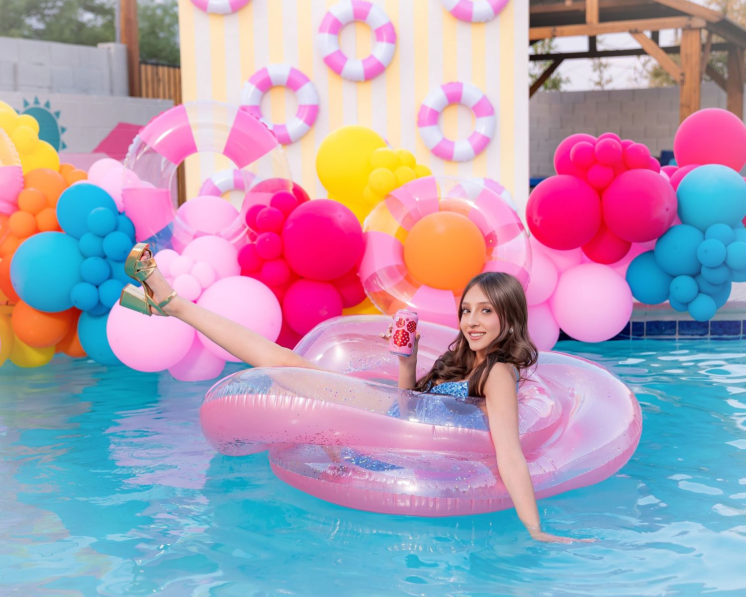 Girl relaxing on pink inflatable ring in pool surrounded by colorful balloons and festive party decorations.