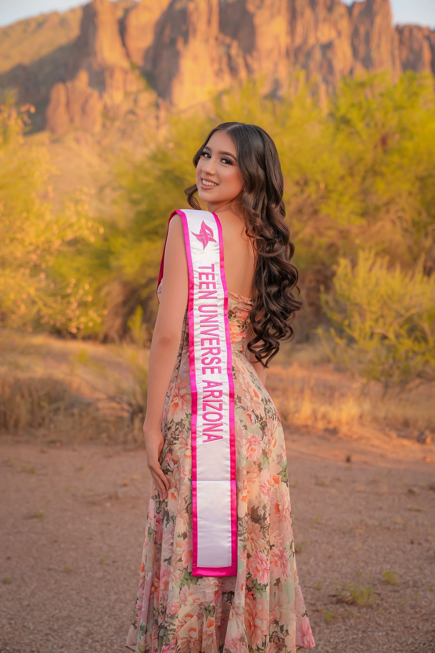 Teen Universe Arizona contestant poses in floral gown with sash, smiling outdoors with desert mountains backdrop.