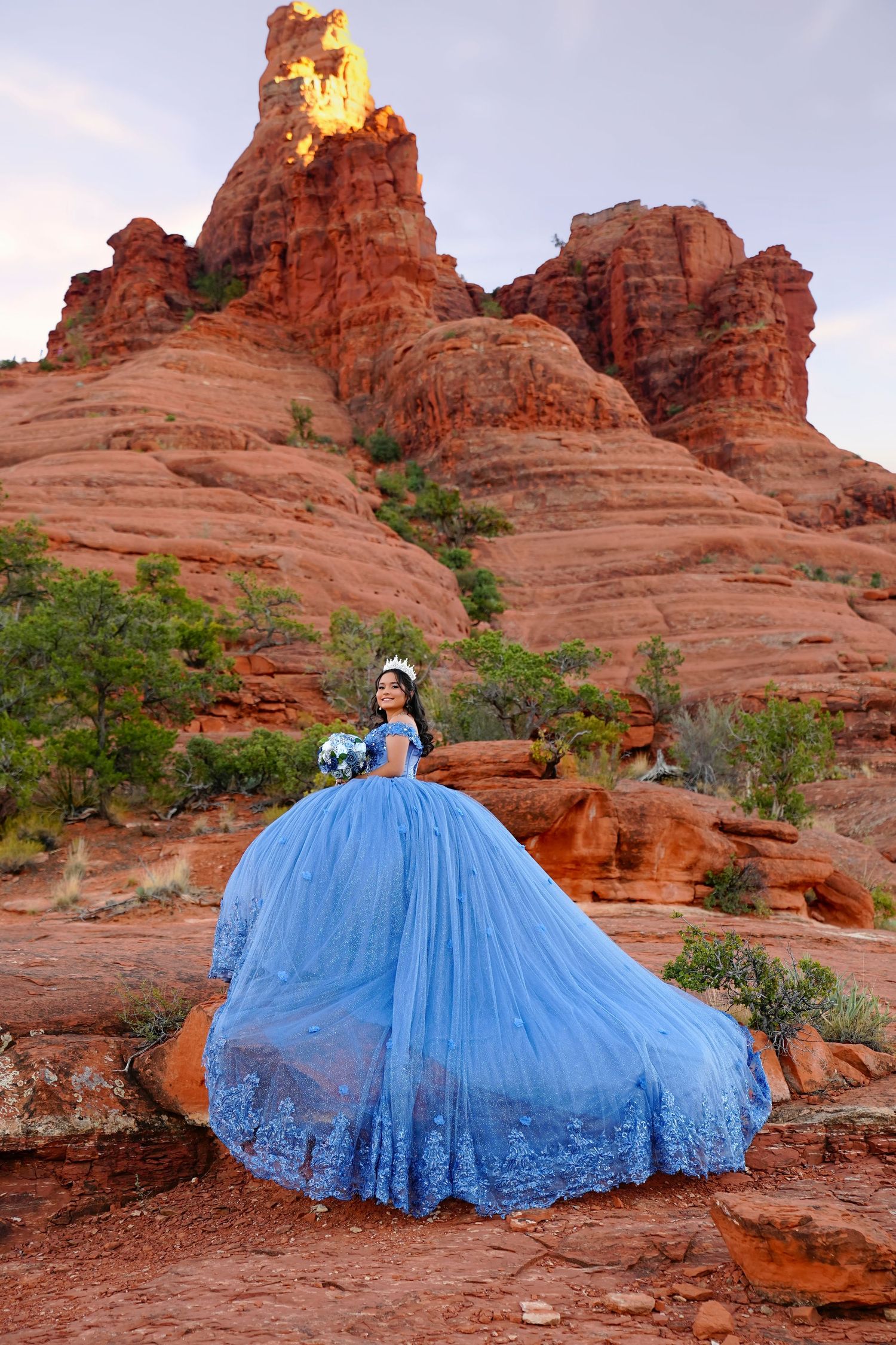 Young woman in a stunning blue ball gown poses among red rock formations in Sedona, Arizona at sunset.