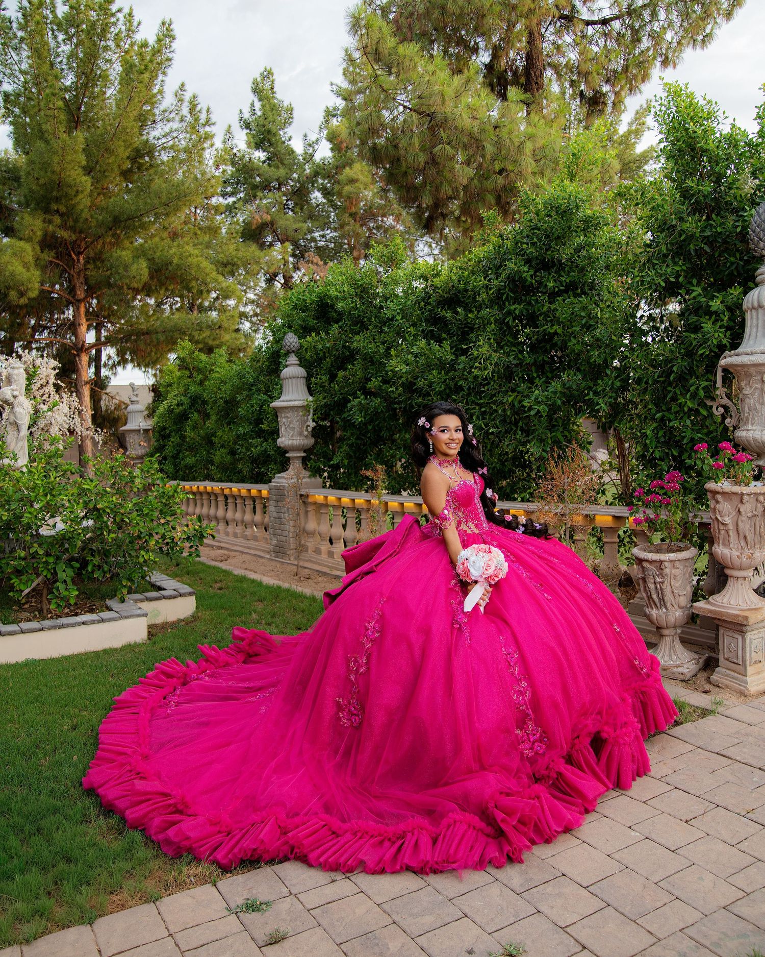 Young woman in a voluminous hot pink quinceañera ball gown with floral details poses in a lush garden.