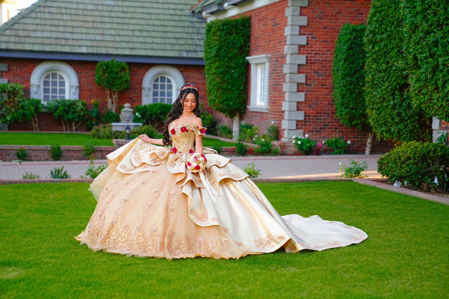 Young woman in an ornate gold quinceañera ball gown with train posing on green lawn by brick estate.
