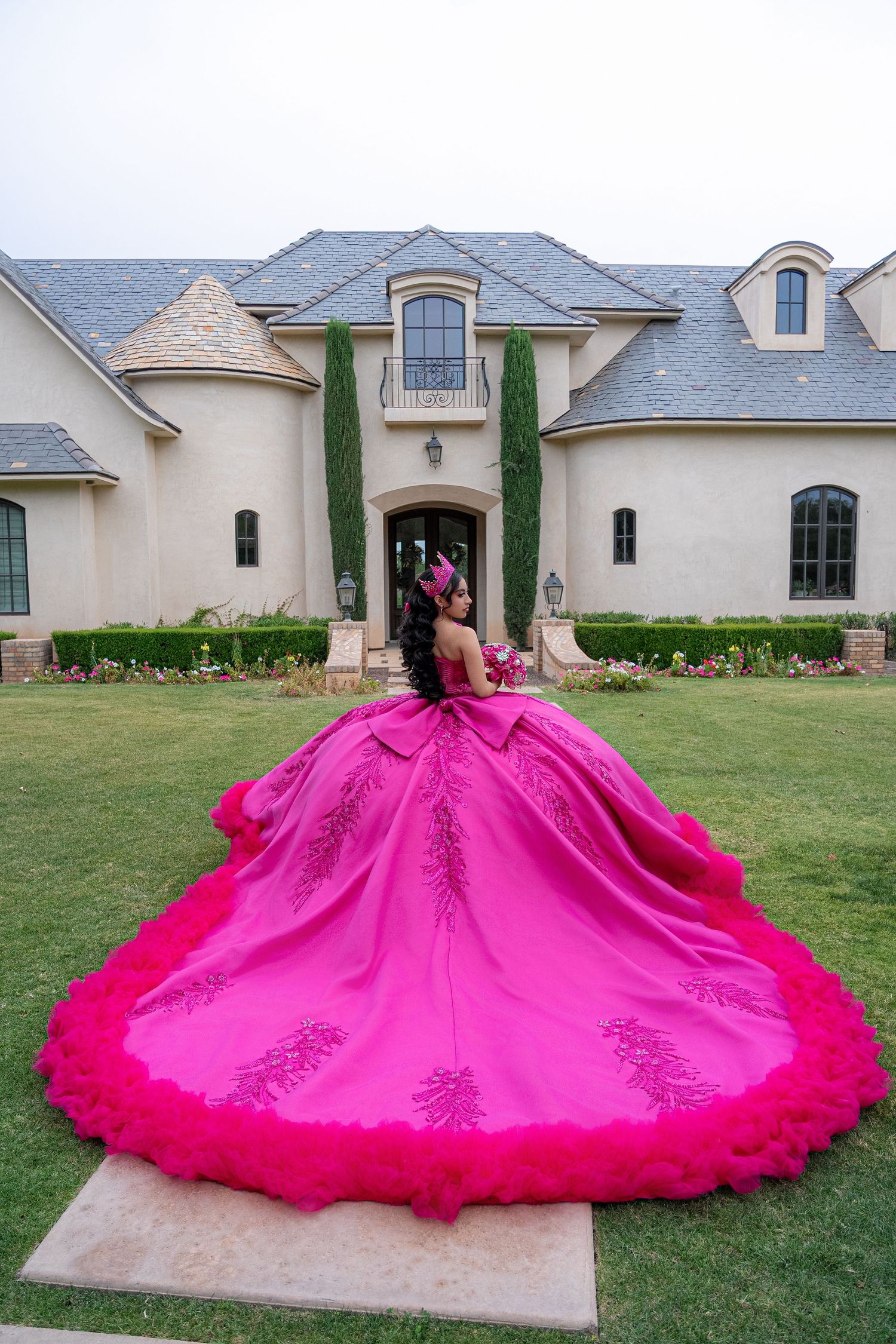 Young woman in stunning hot pink quinceañera ball gown with floral embroidery poses in front of elegant mansion.