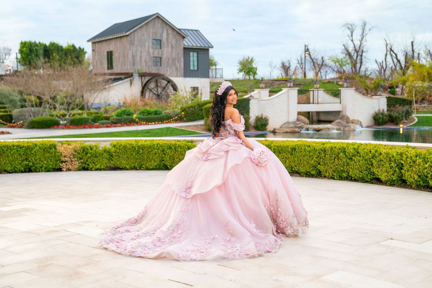 Young woman in a pink ball gown quinceañera dress poses gracefully on a stone patio with lush gardens.
