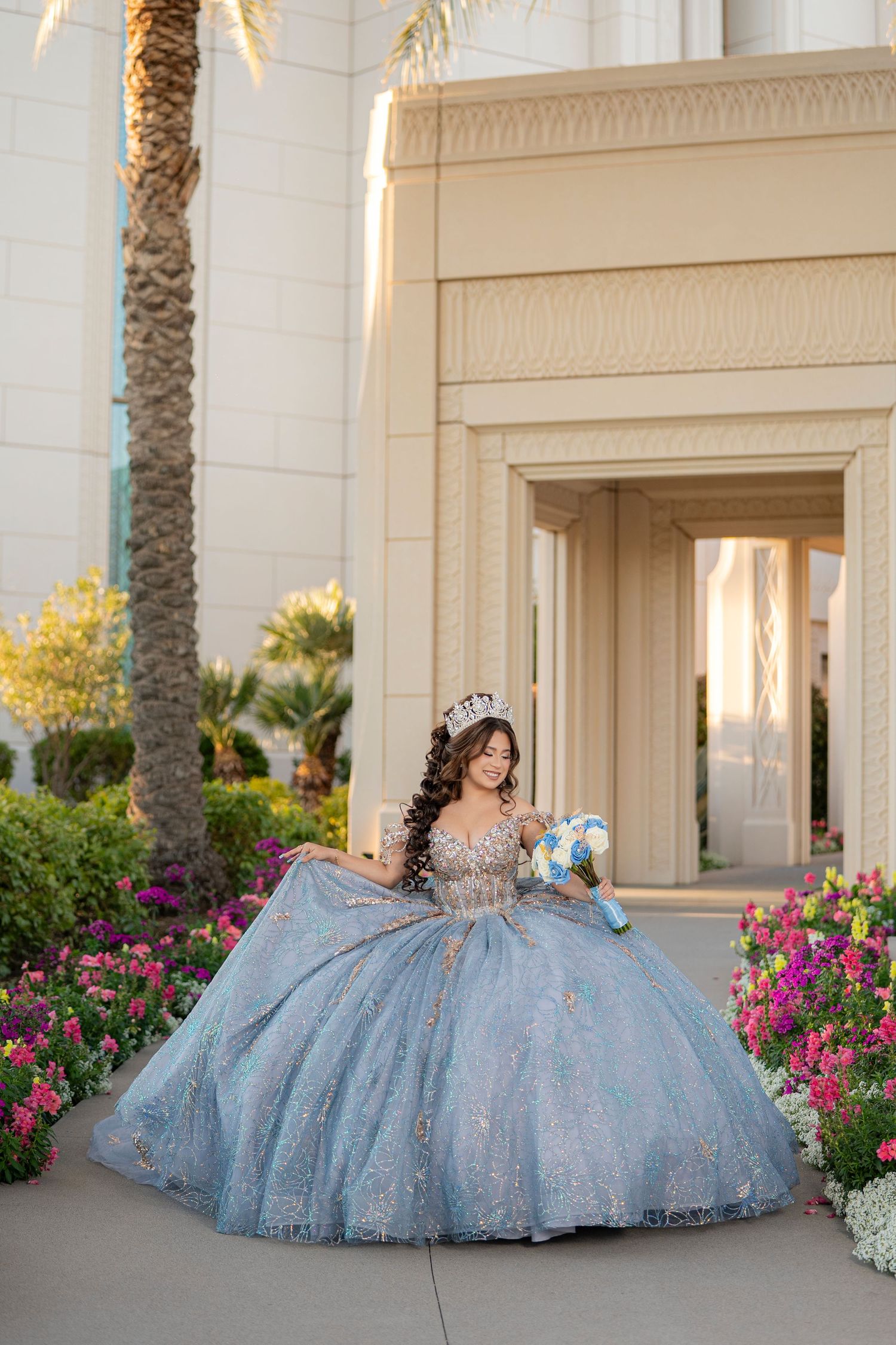 Young woman in blue quinceañera ball gown with crown holds bouquet beside colorful garden and elegant building.