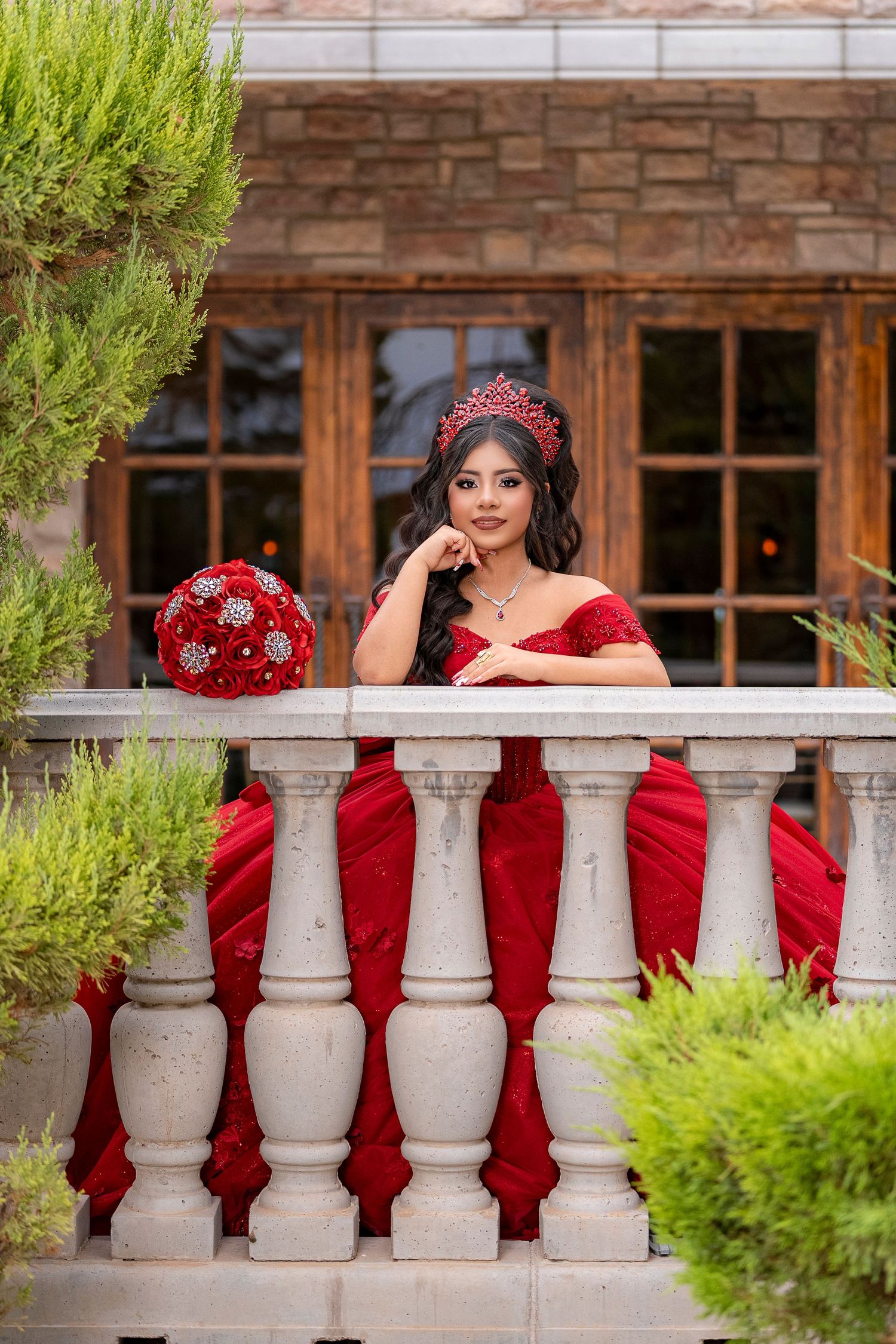 Young woman in red off-shoulder quinceañera gown and crown poses gracefully on stone balcony with matching red bouquet.