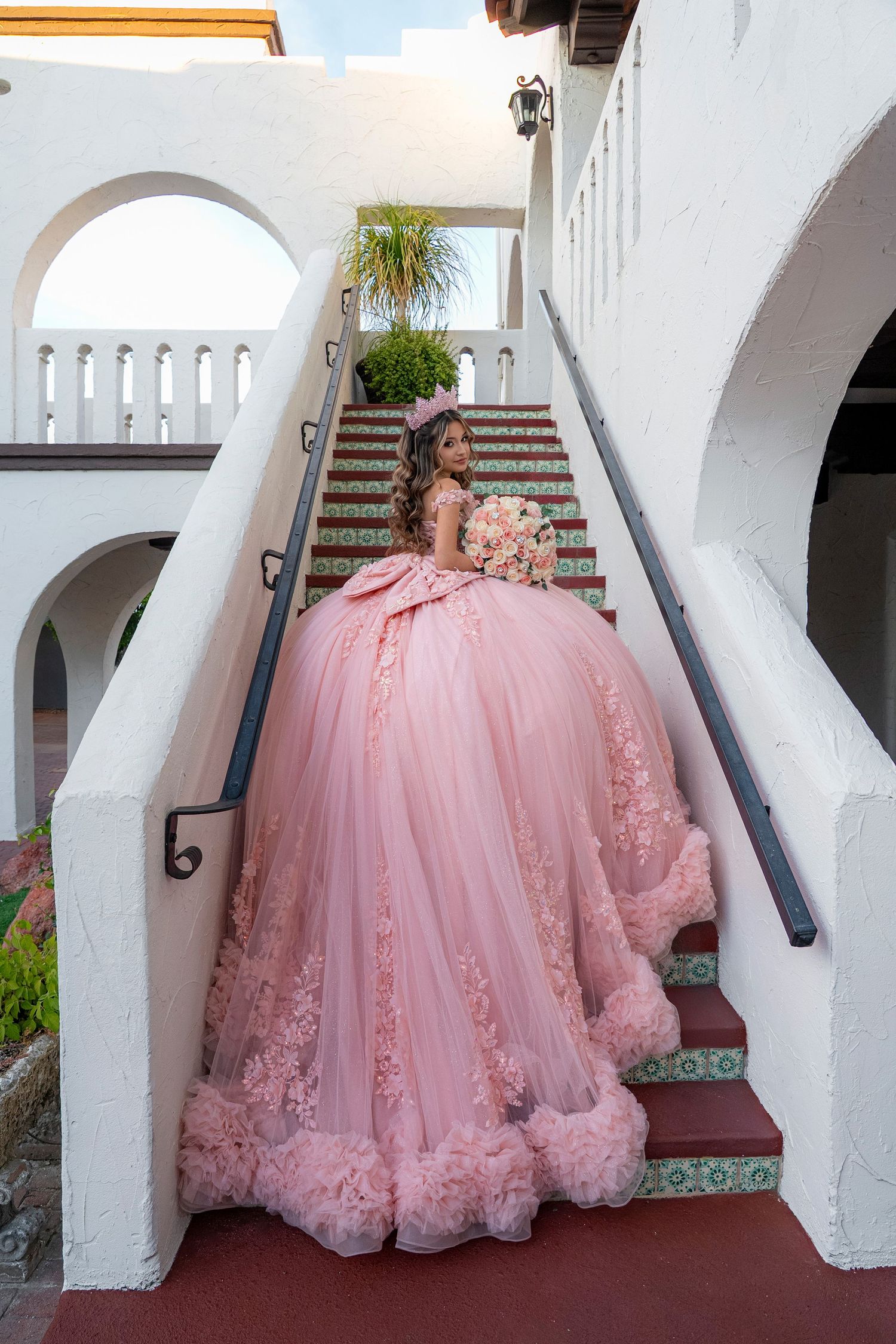 Young woman in elegant pink quinceañera ball gown with floral details poses on decorative tiled stairs.