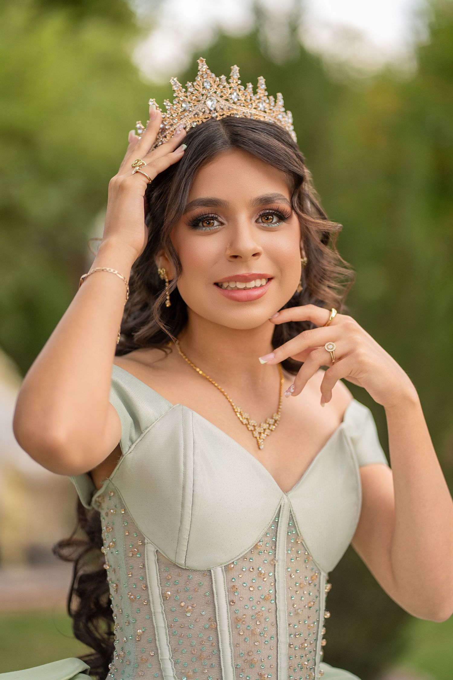 Young woman in mint green gown wearing gold crown and jewelry, smiling outdoors at a quinceañera celebration.