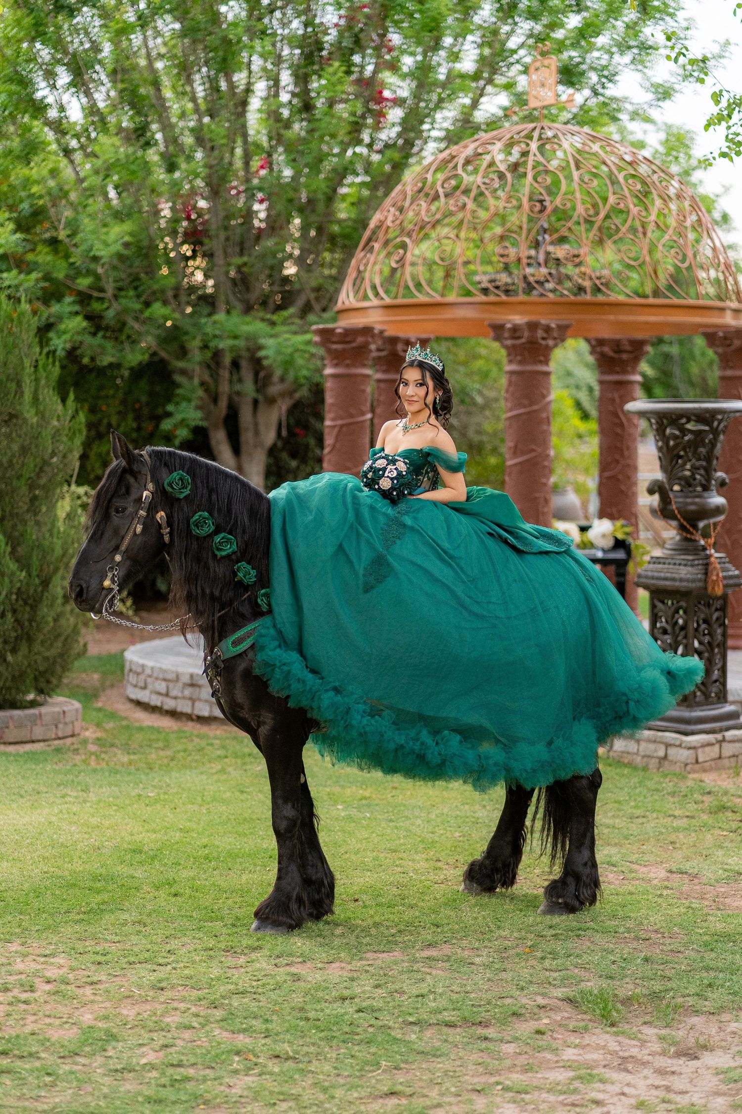Young woman in emerald green quinceañera gown riding a black horse in a garden with ornate gazebo.
