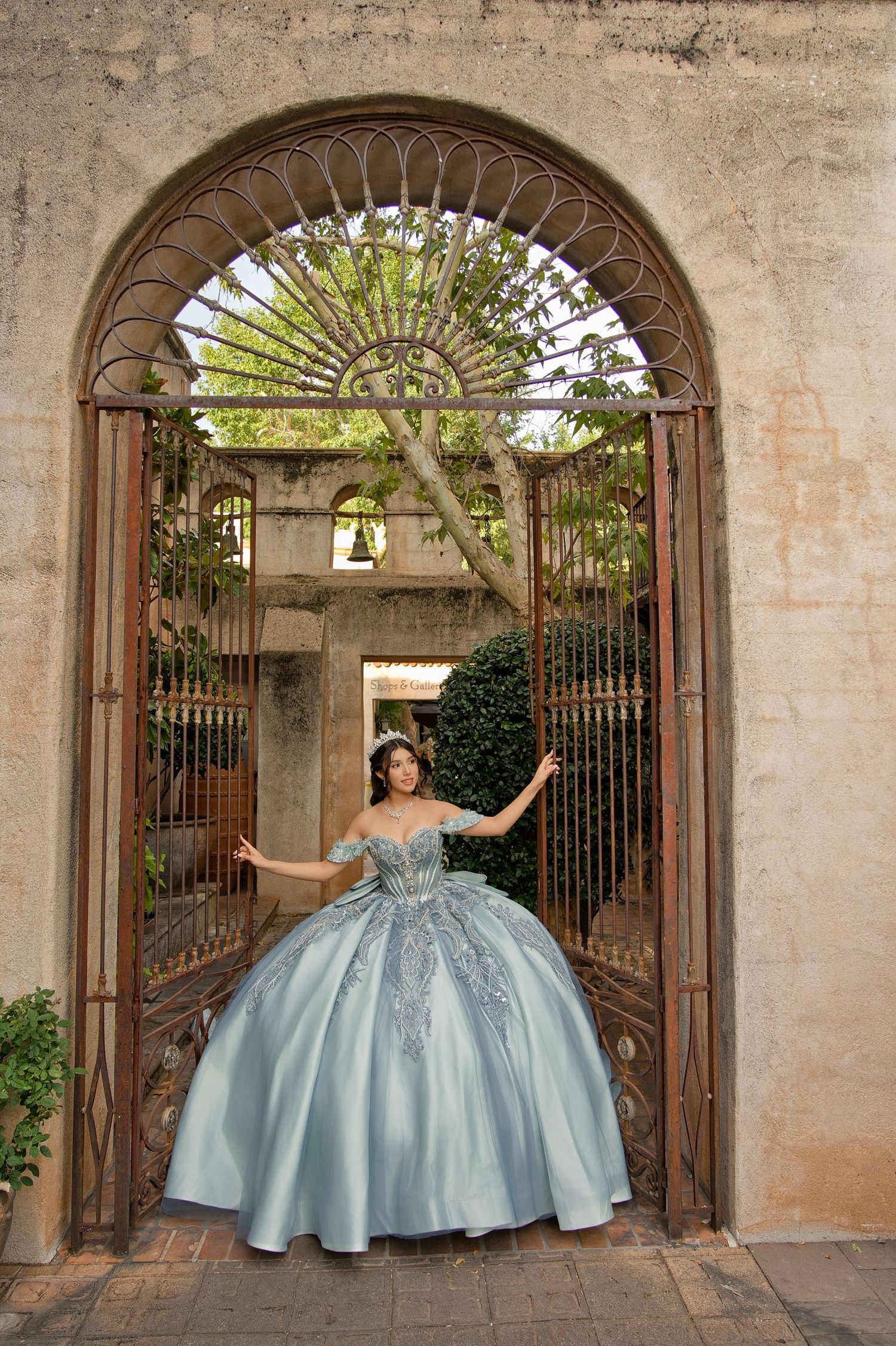 Young woman in a powder blue off-shoulder quinceañera ball gown poses in an ornate iron garden gate archway.