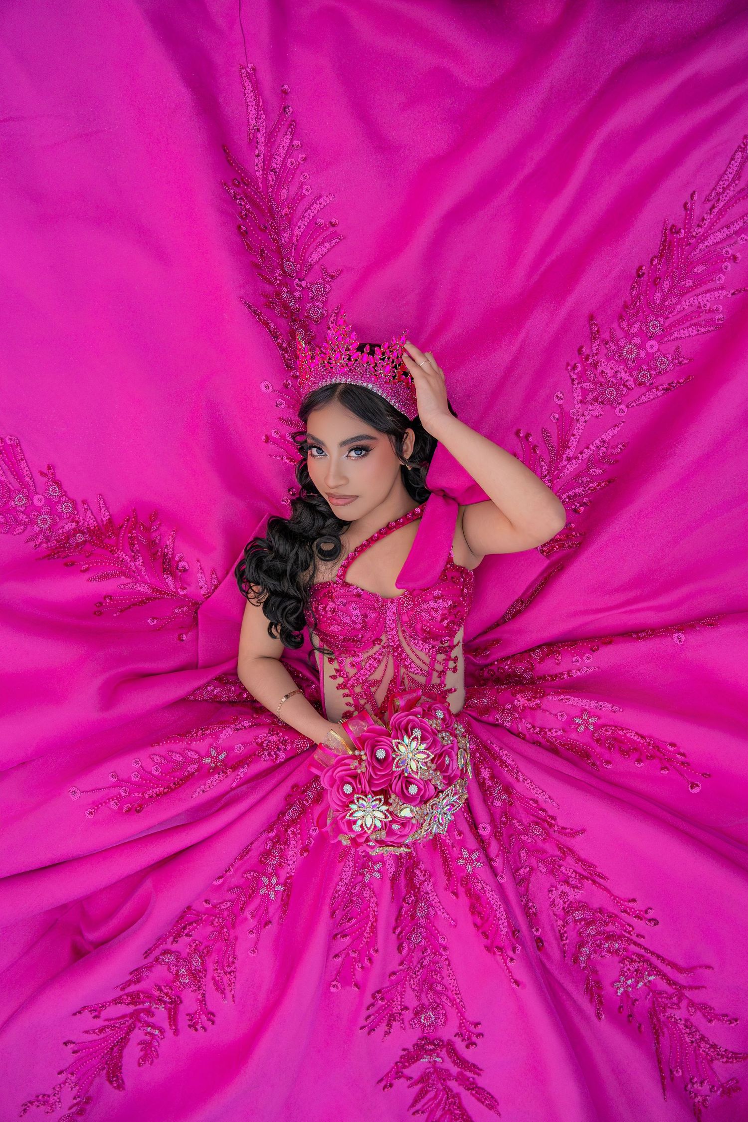 Young woman in elaborate hot pink quinceañera ball gown with crown lies surrounded by flowing fabric.