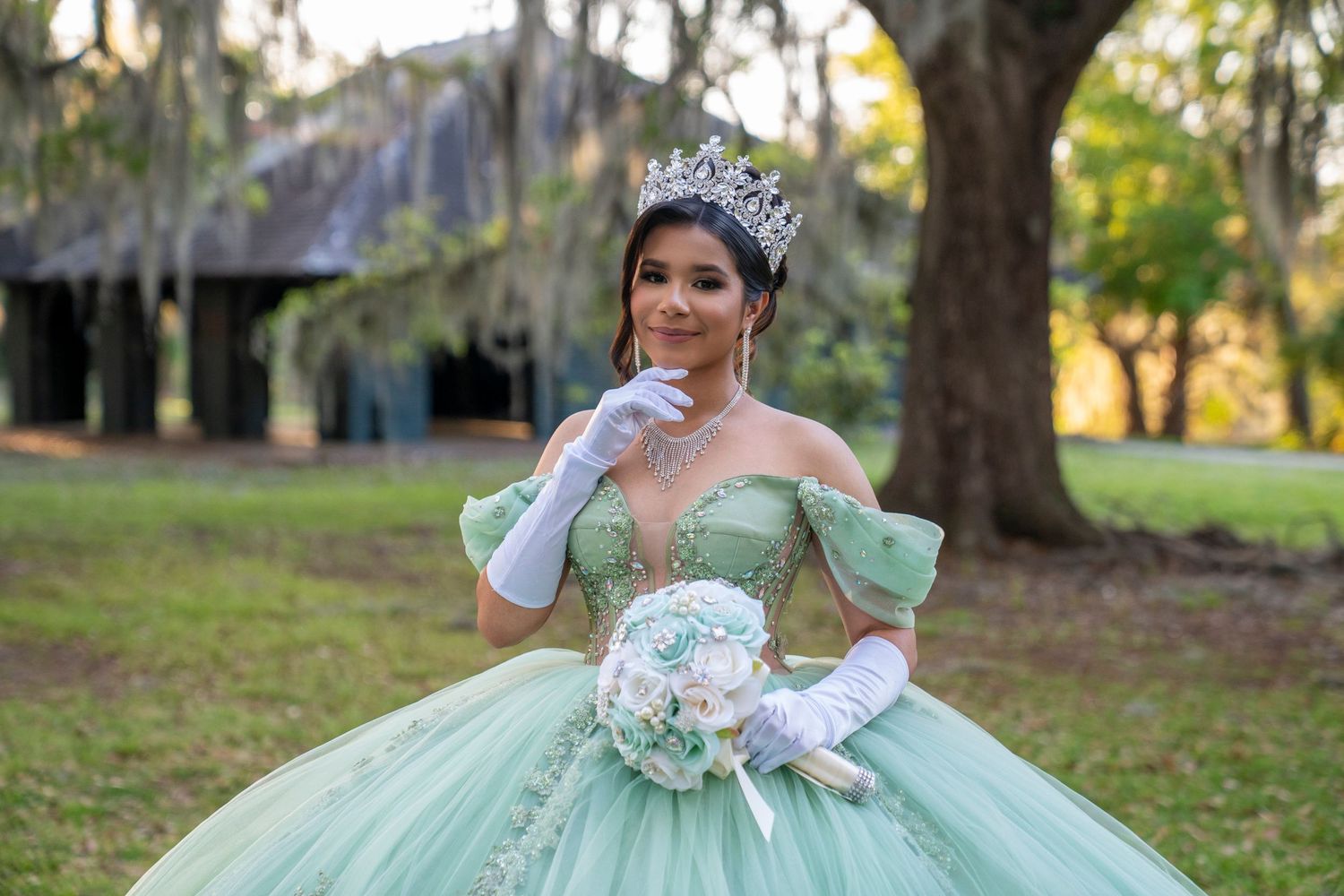 Young woman in mint green quinceañera ball gown with crown and white gloves holding floral bouquet outdoors.