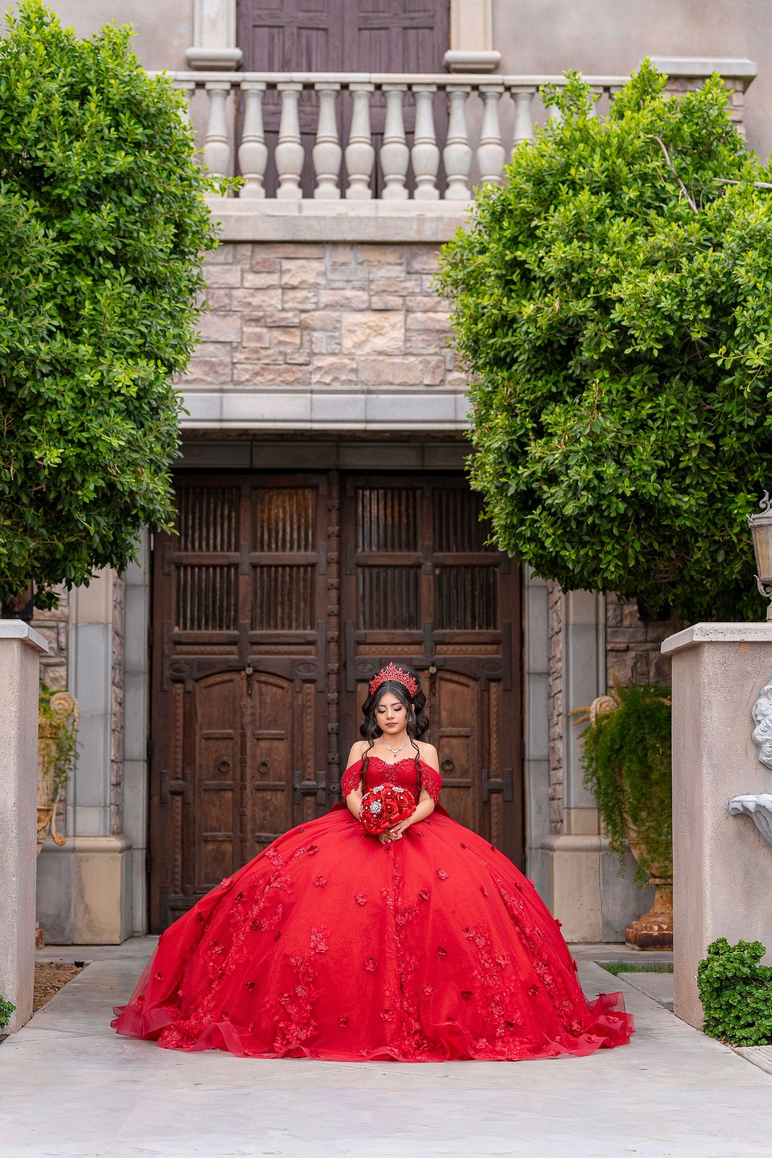 Young woman in a red ball gown quinceañera dress poses before ornate wooden doors flanked by lush green trees.
