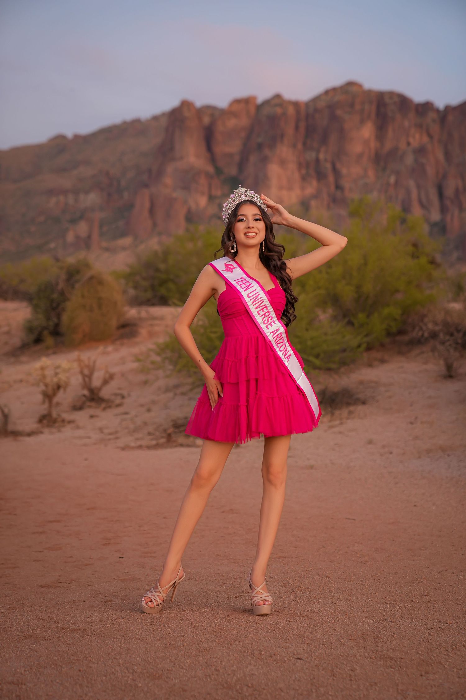 Young woman in pink dress and tiara wearing a pageant sash poses in desert landscape with rocky mountains at sunset.