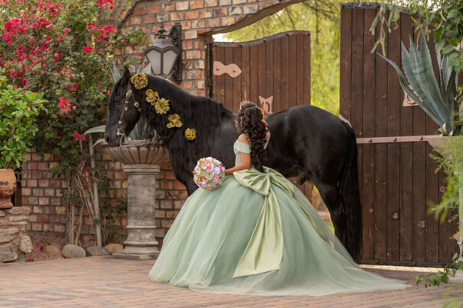 Young woman in sage green quinceañera gown poses with black horse adorned with yellow flowers at rustic brick venue.