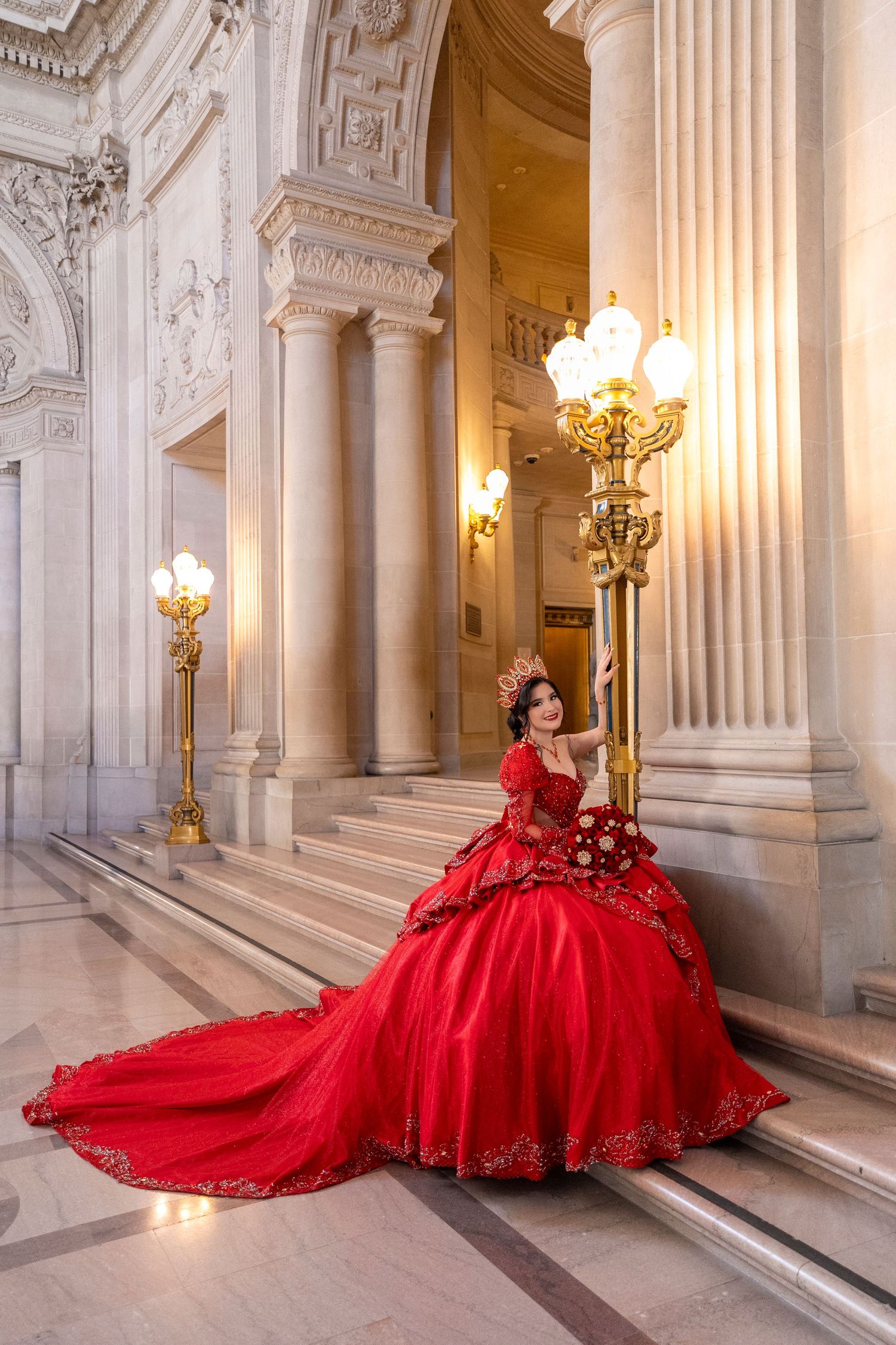 Elegant woman in red quinceañera gown sits beside ornate gold lamp post among classical marble columns.
