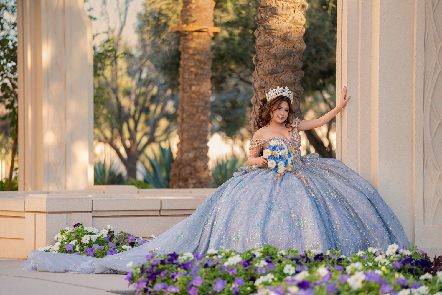 Young woman in a blue quinceañera ball gown with tiara poses among purple and white flowers at an elegant venue.