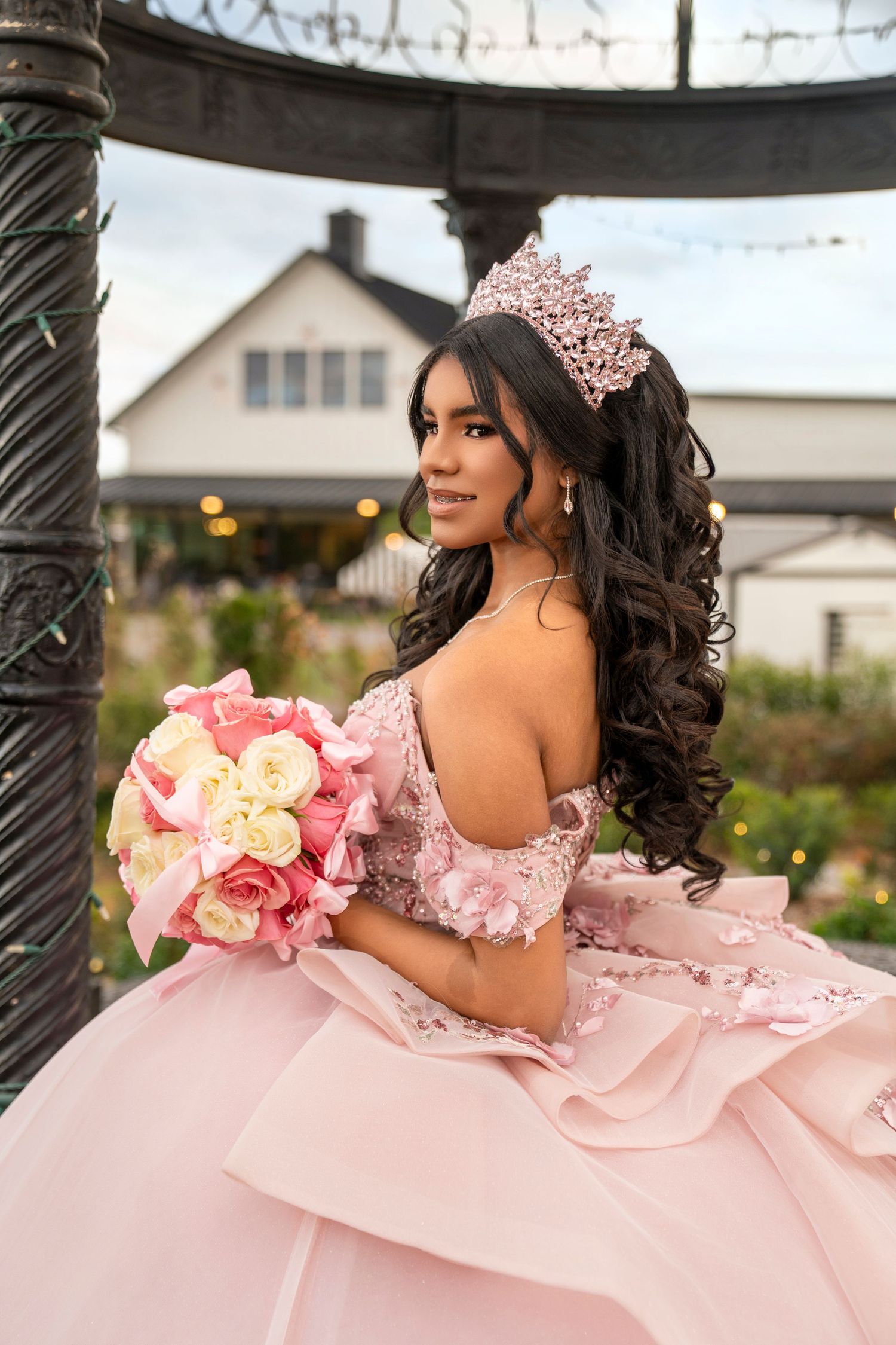 Young woman in pink quinceañera gown and crown holds floral bouquet outdoors near decorative iron gazebo.