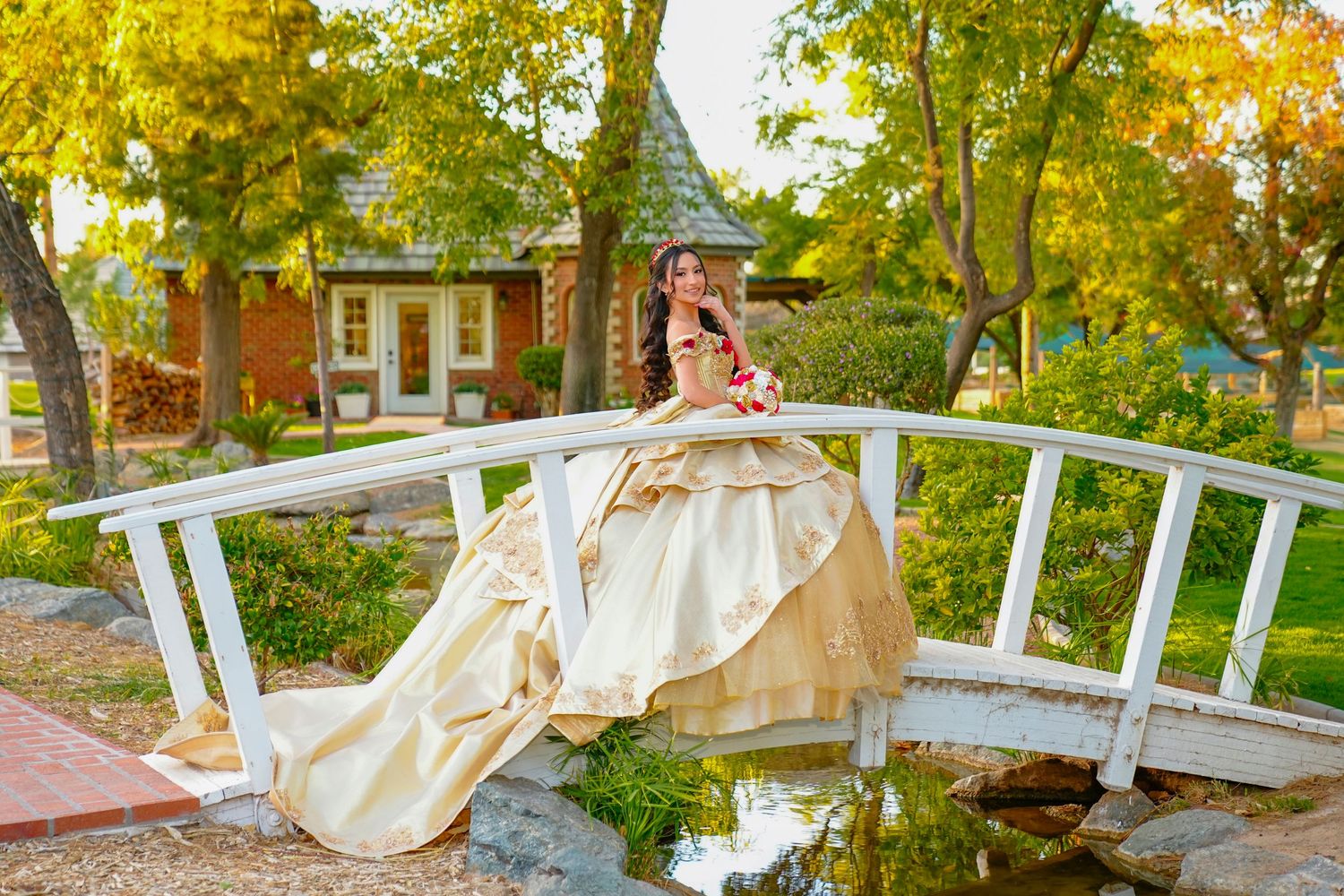 Young woman in a gold quinceañera ball gown posing gracefully on a white arched garden bridge.