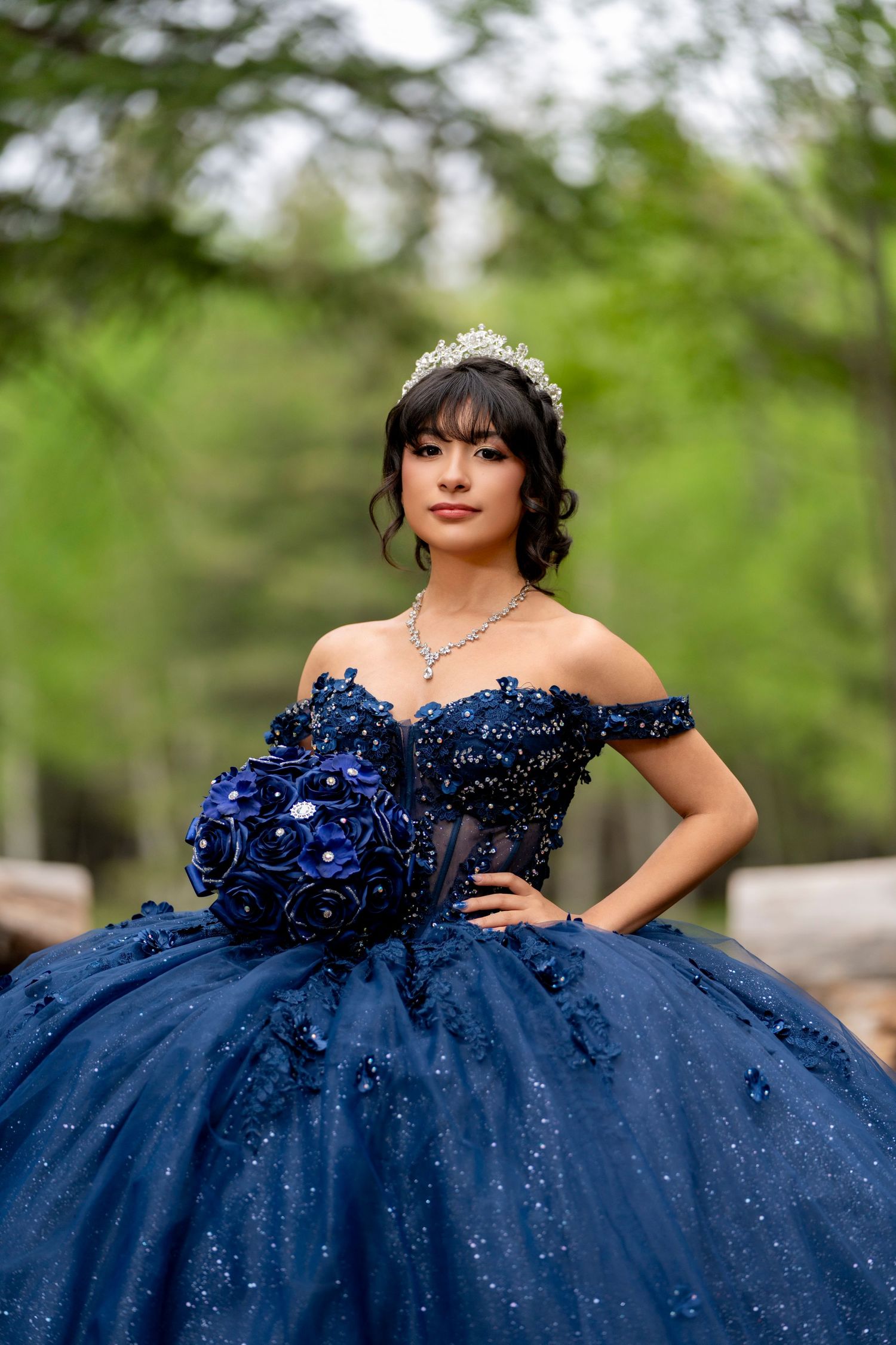 Young woman in navy blue quinceañera ball gown with tiara, holding floral bouquet outdoors.