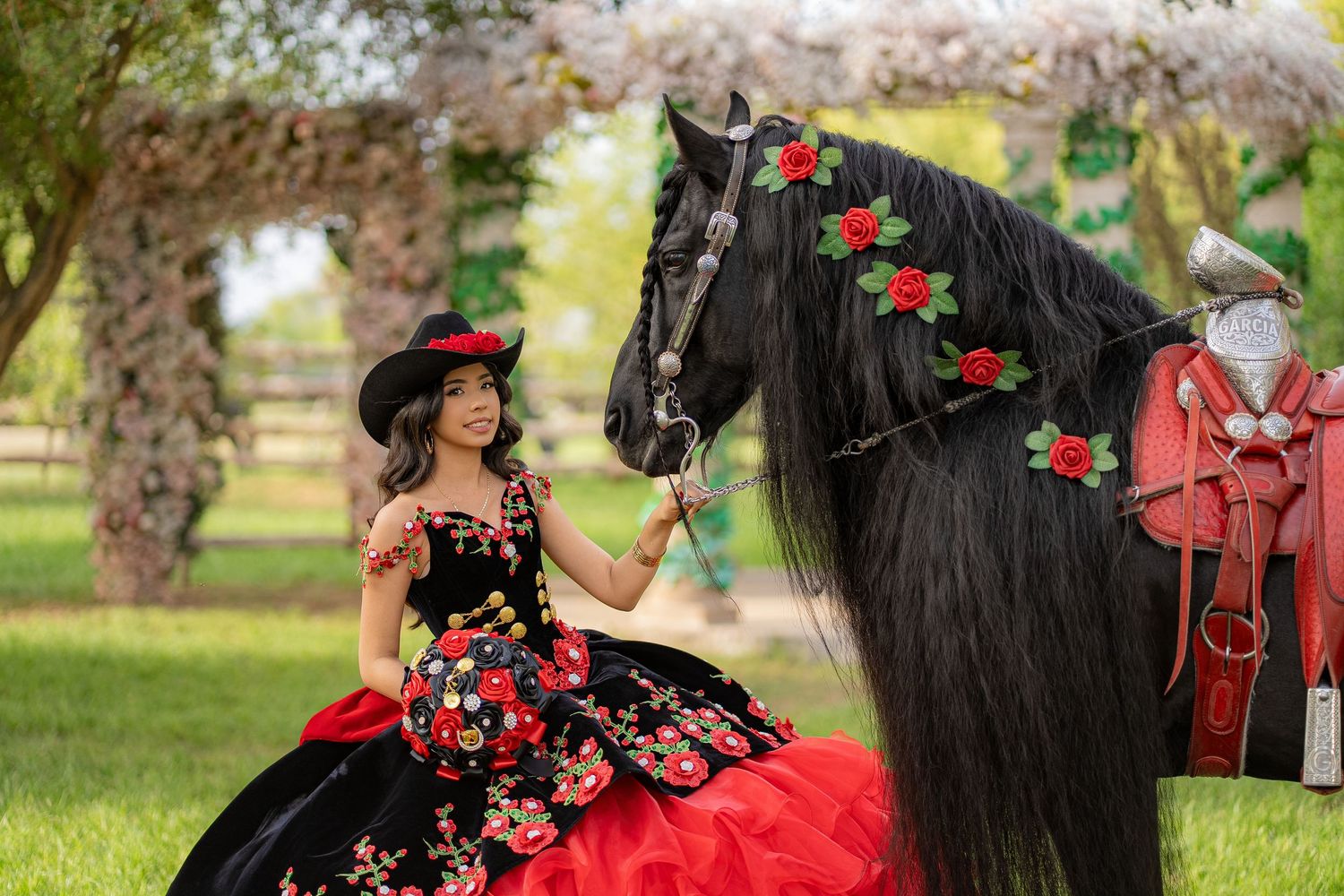Young woman in red floral quinceañera gown and hat poses with decorated black horse outdoors.