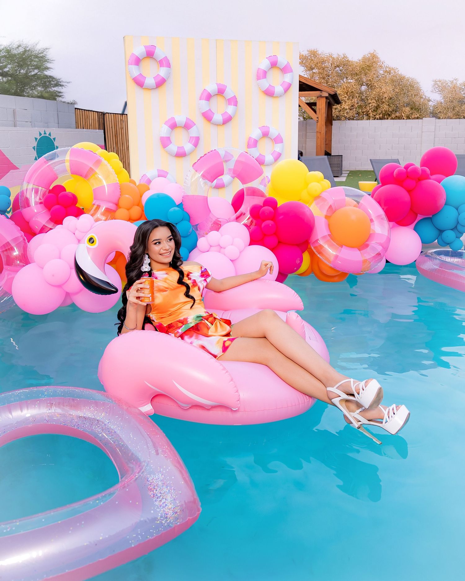 Woman relaxing on pink flamingo pool float surrounded by colorful balloons and pool toys at a festive summer party.