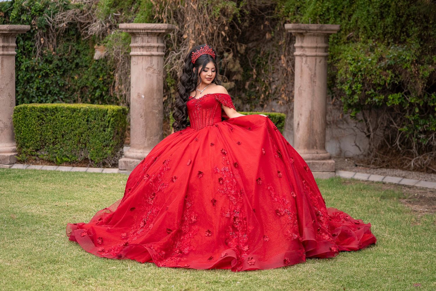 Young woman in a stunning red off-shoulder ball gown with floral details poses outdoors near stone columns.
